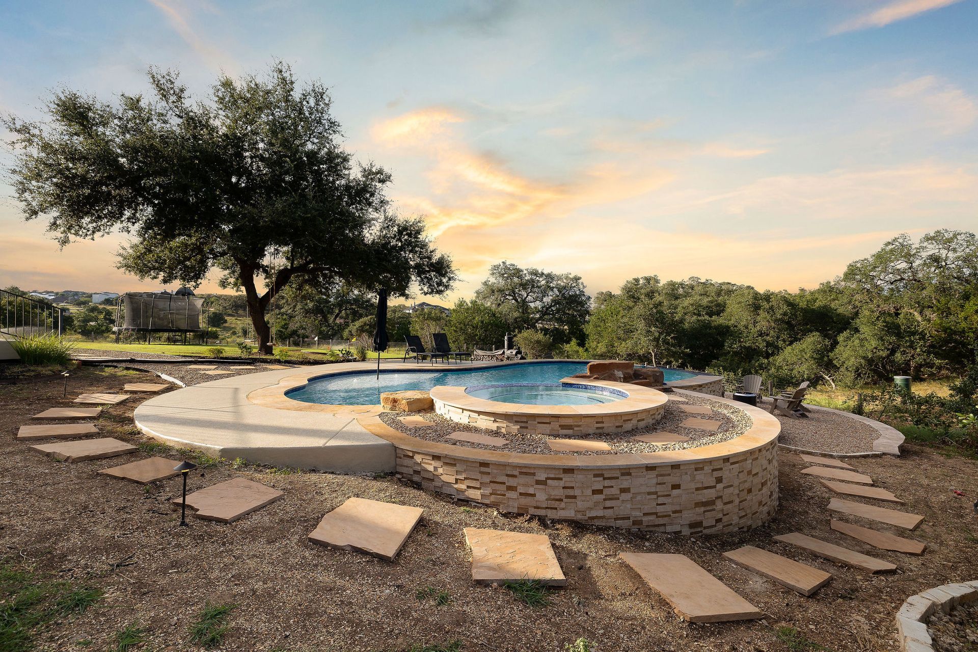 Circular stone fire pit with a blue basin, surrounded by stepping stones in a landscaped yard at sunset