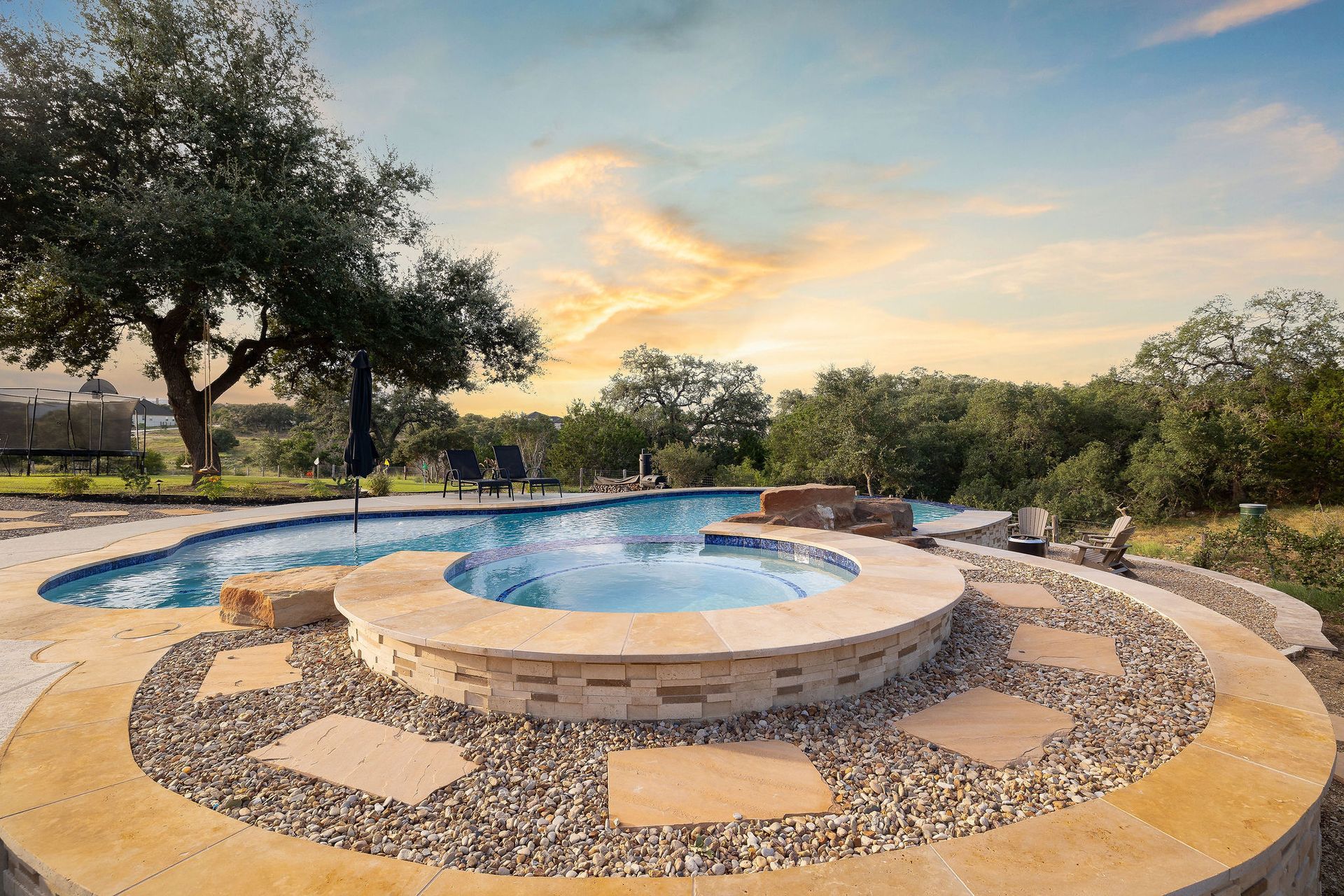 Circular stone patio with a hot tub beside a swimming pool at sunset, surrounded by trees and lawn
