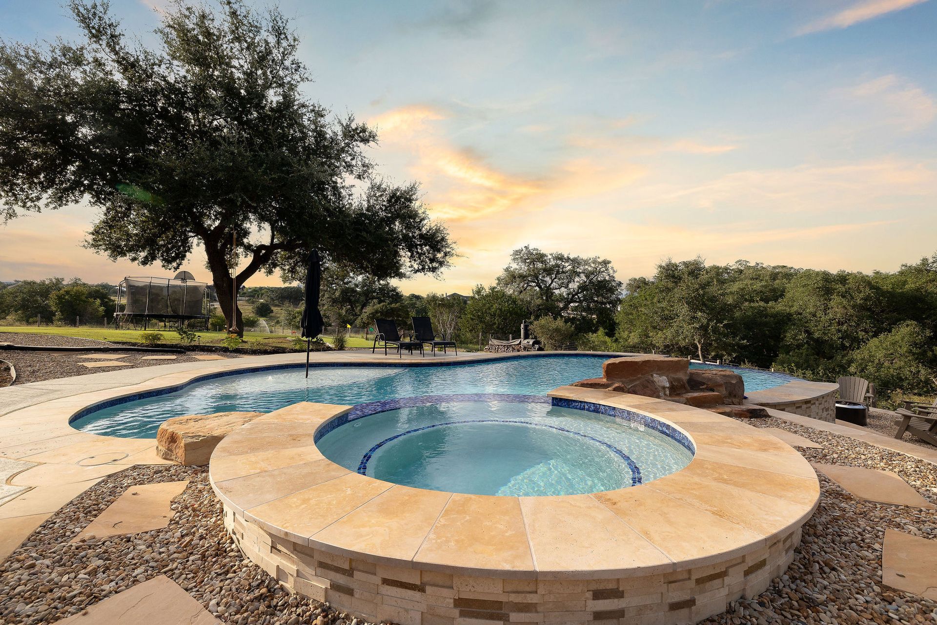 Backyard pool and hot tub at sunset with stone patio and trees