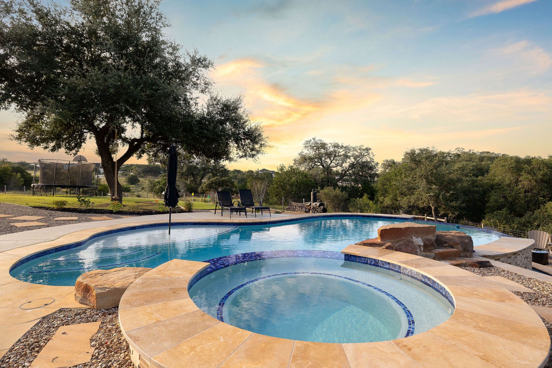 Backyard pool with a raised spa, stone deck, and trees at sunset