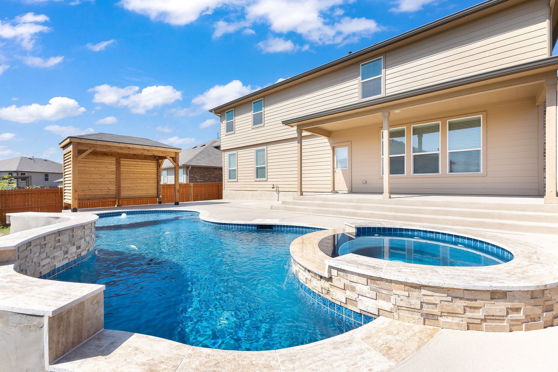 Backyard with a blue pool, round hot tub, and beige house under a bright sky