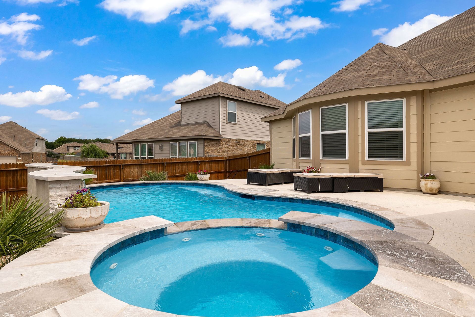 Backyard with blue swimming pool and hot tub beside a beige house under a partly cloudy sky