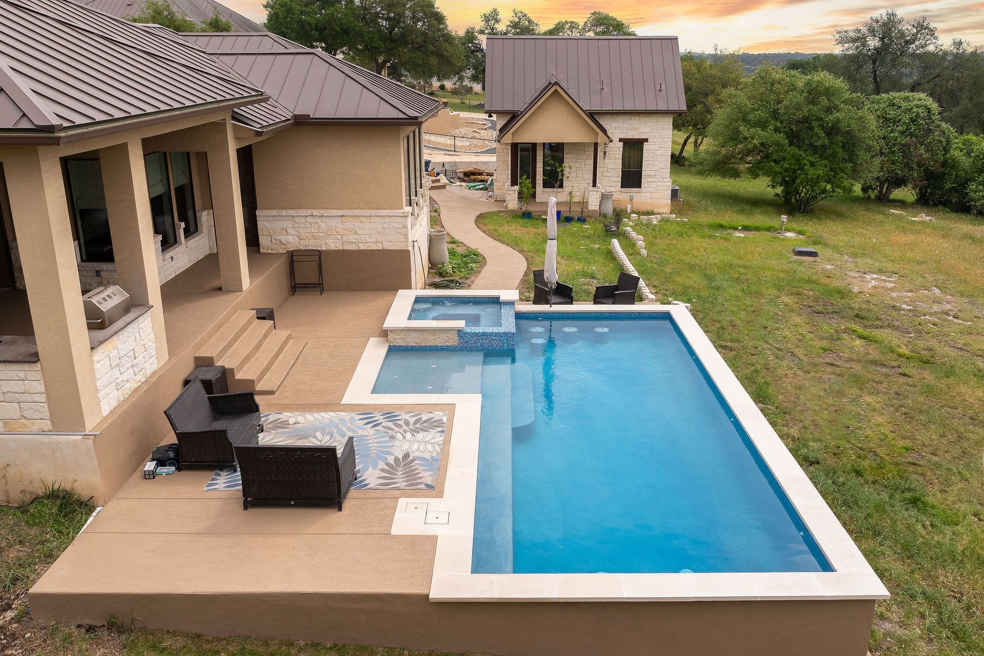 Modern house with a blue swimming pool and patio seating, overlooking a green backyard at sunset