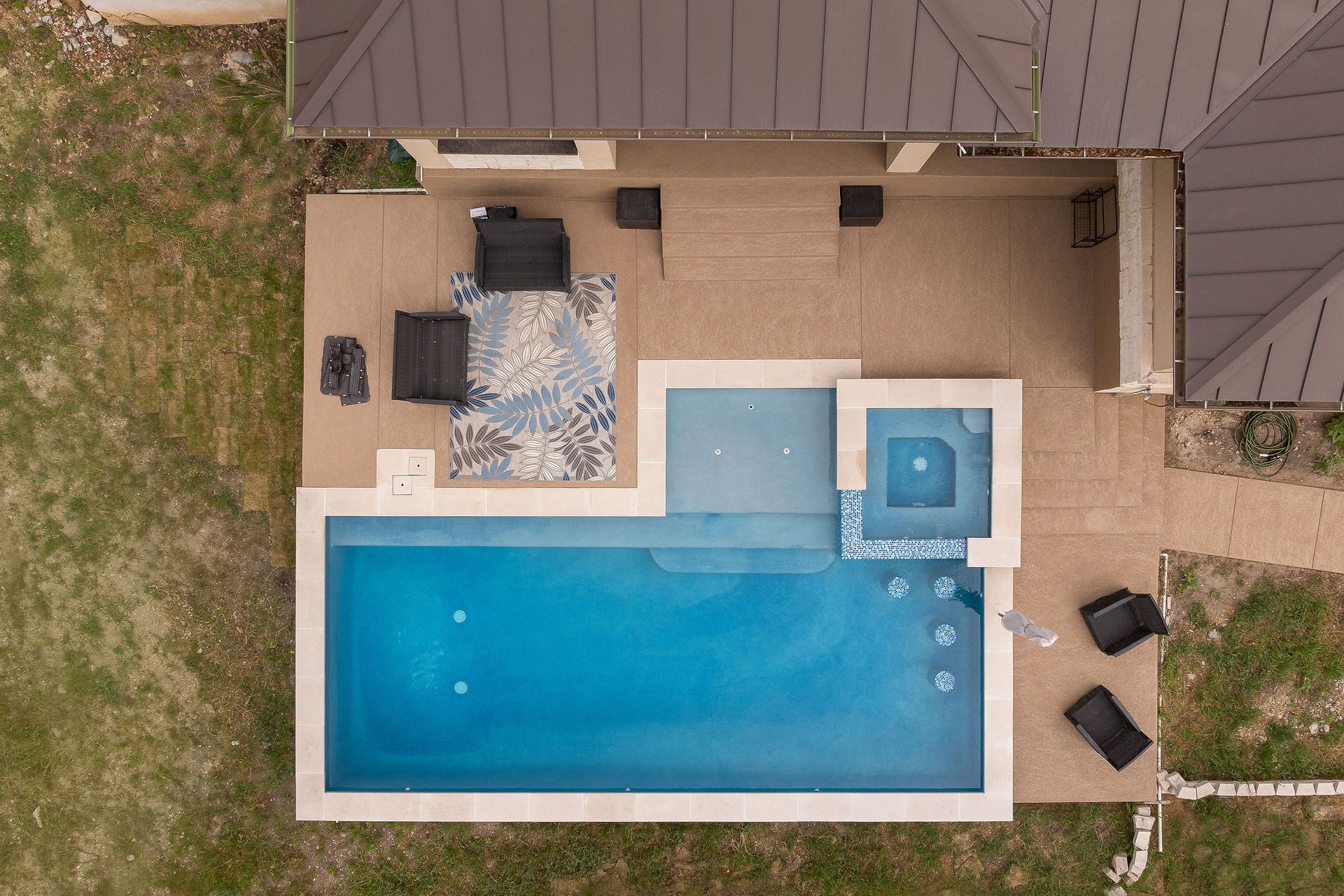 Overhead view of a backyard pool and patio with lounge chairs and a dining table beside a house