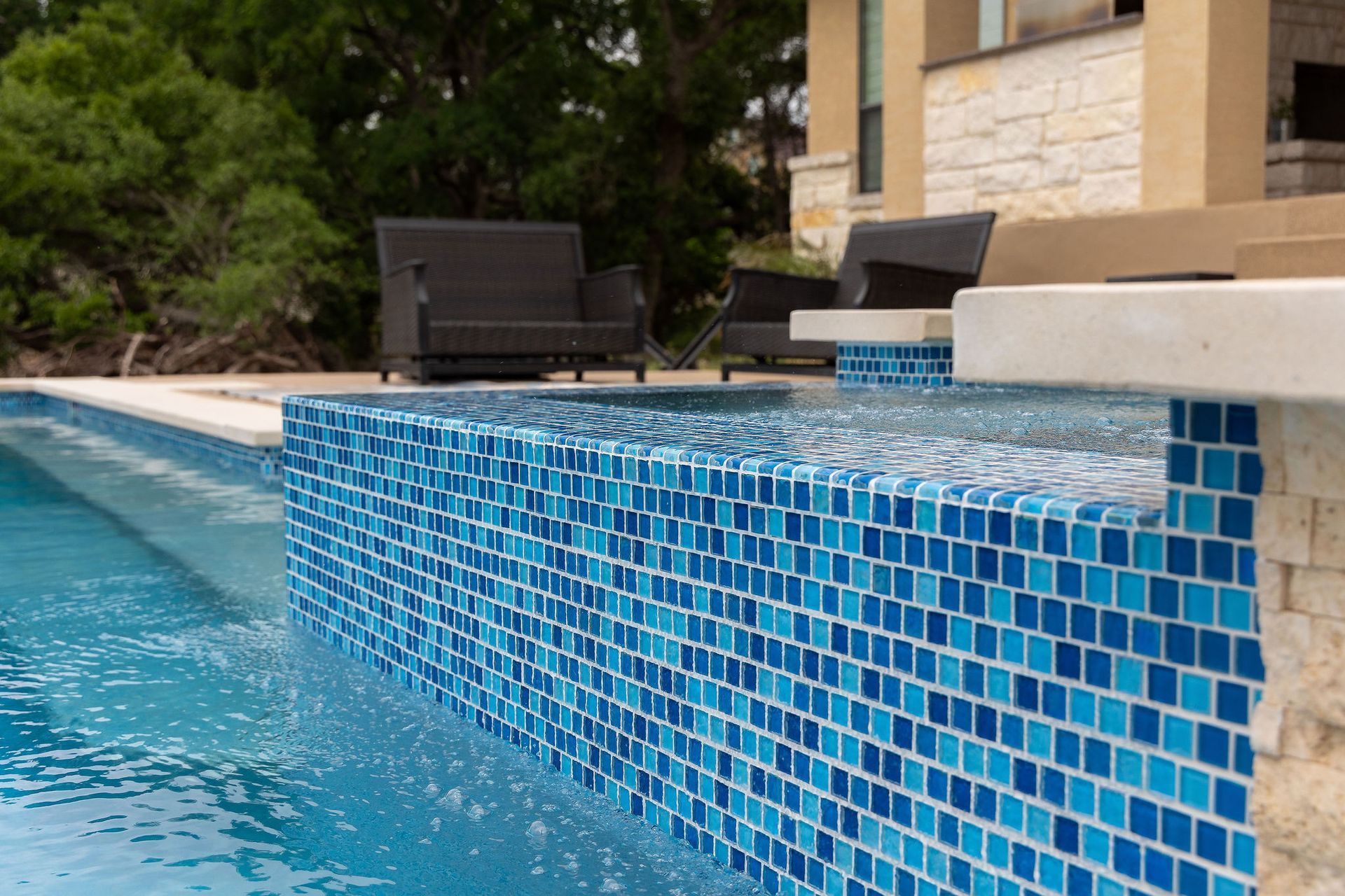 Blue-tiled pool edge beside a patio and lounge chairs, with trees and a house in the background