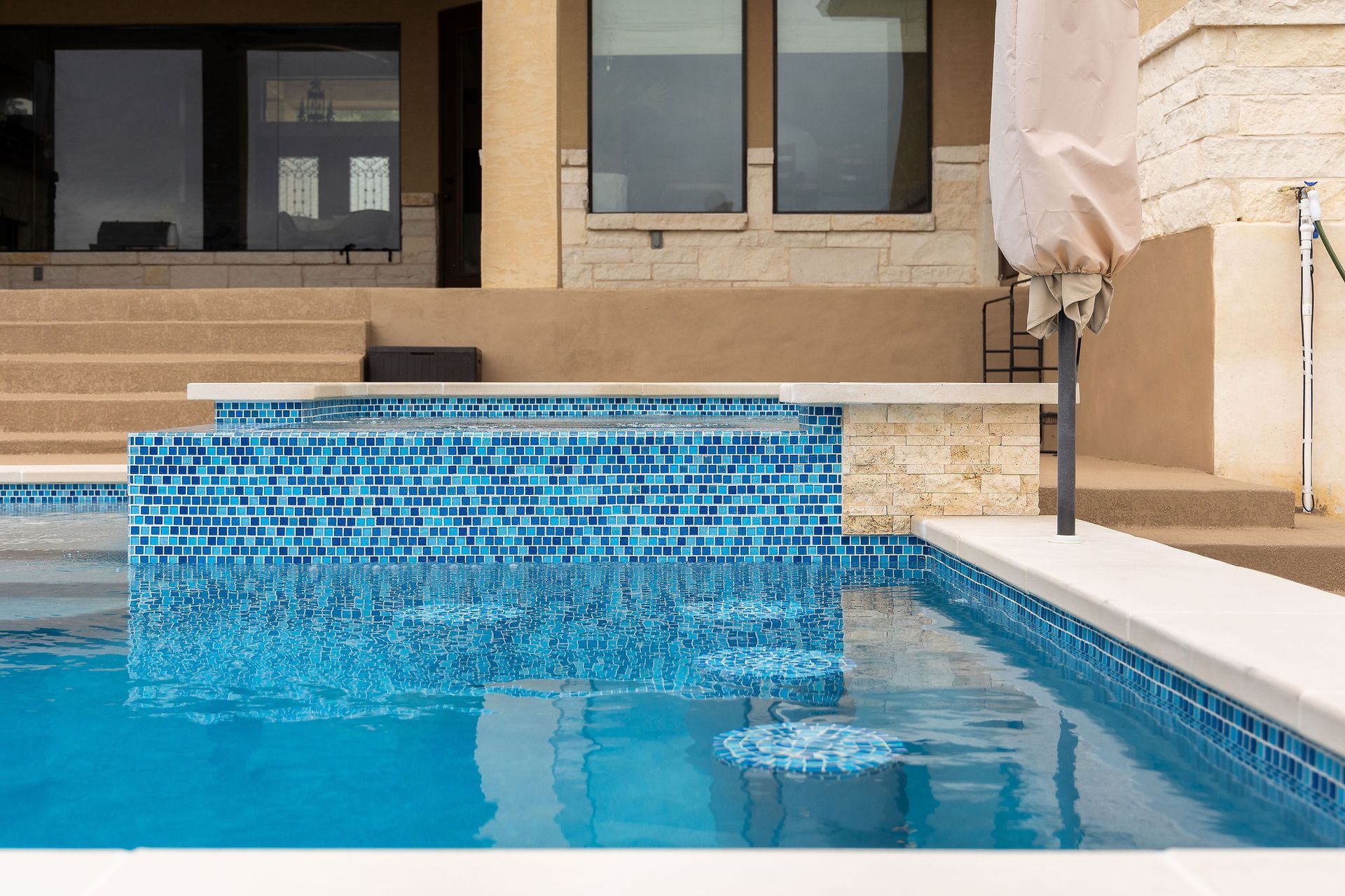 Blue-tiled swimming pool with a raised spillover wall beside a beige building and patio chair