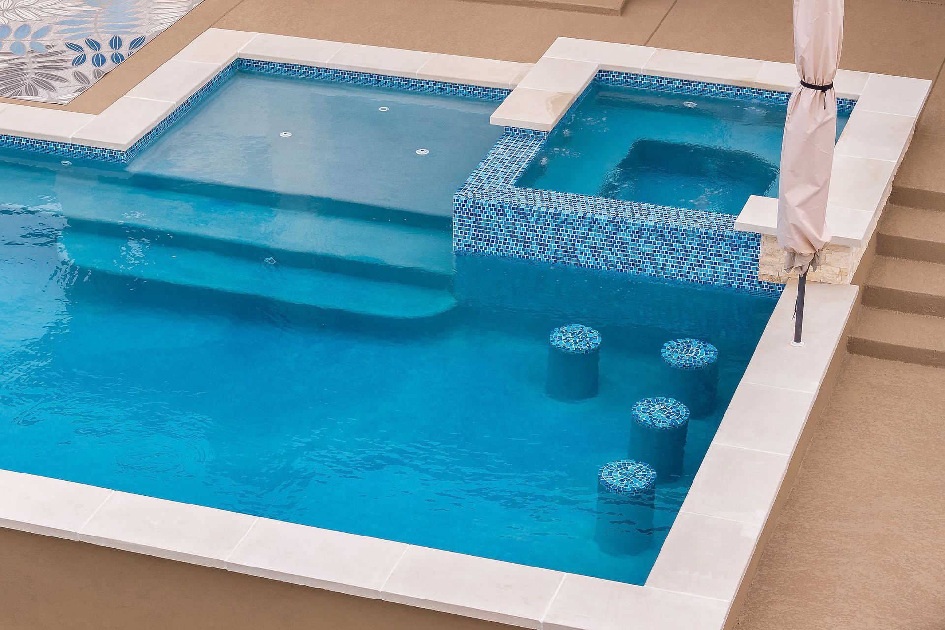 Blue swimming pool with steps, water feature, and covered seating area beside a tiled deck