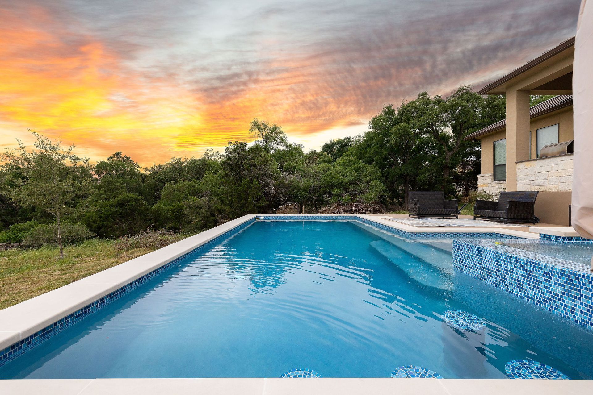 Backyard pool at sunset with blue water, white deck, and trees beside a modern house