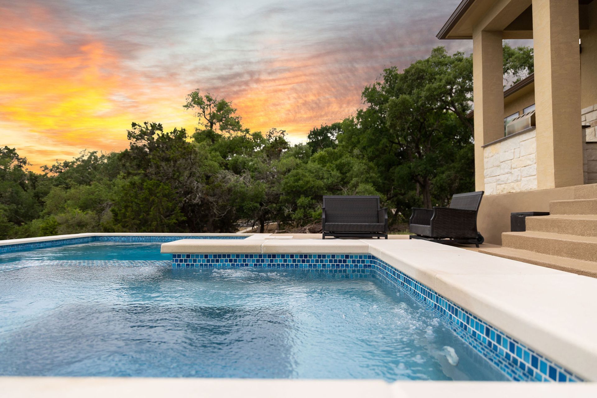 Backyard pool at sunset with blue water, lounge chairs, and trees under a colorful sky