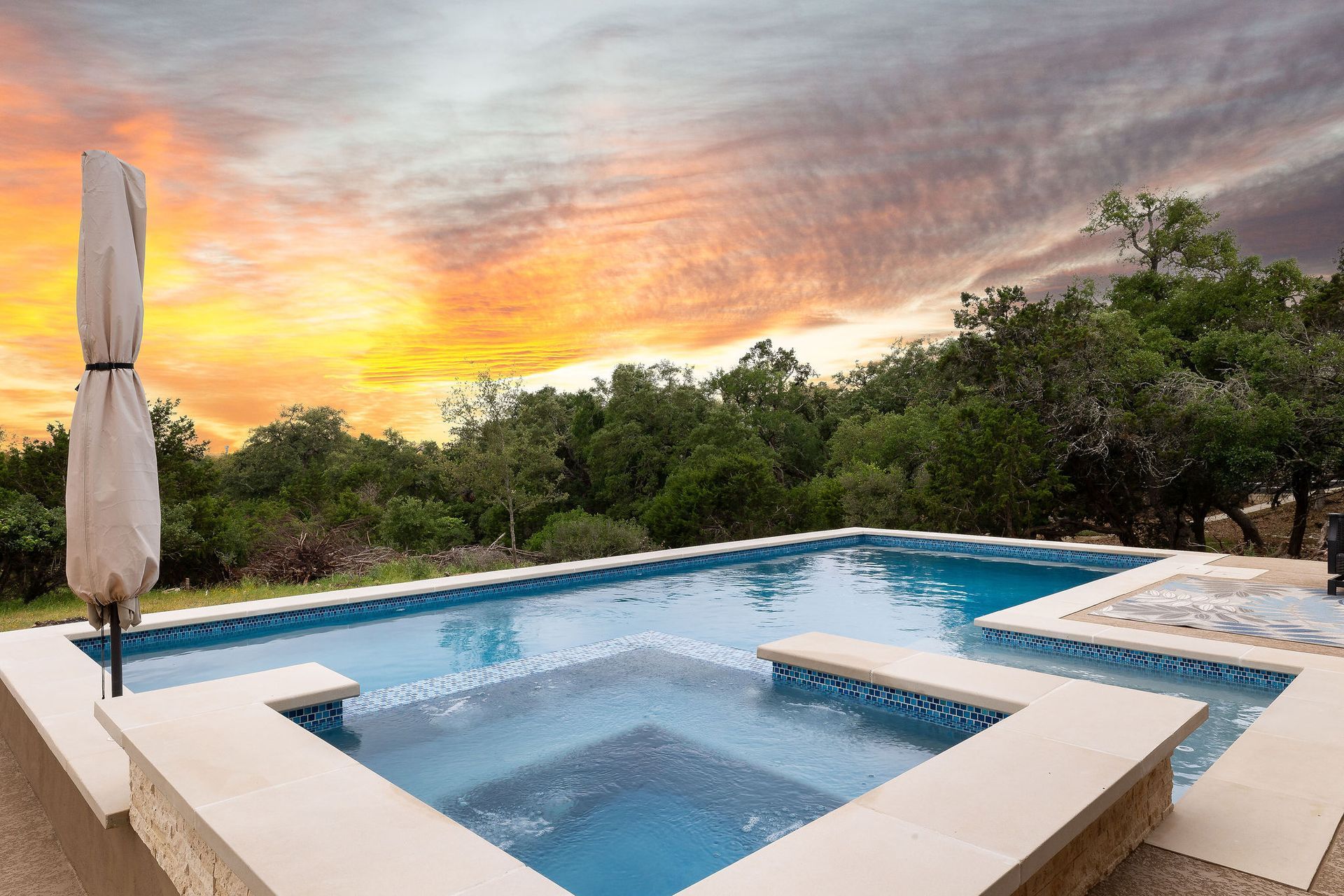Infinity pool and hot tub at sunset with trees and a closed patio umbrella