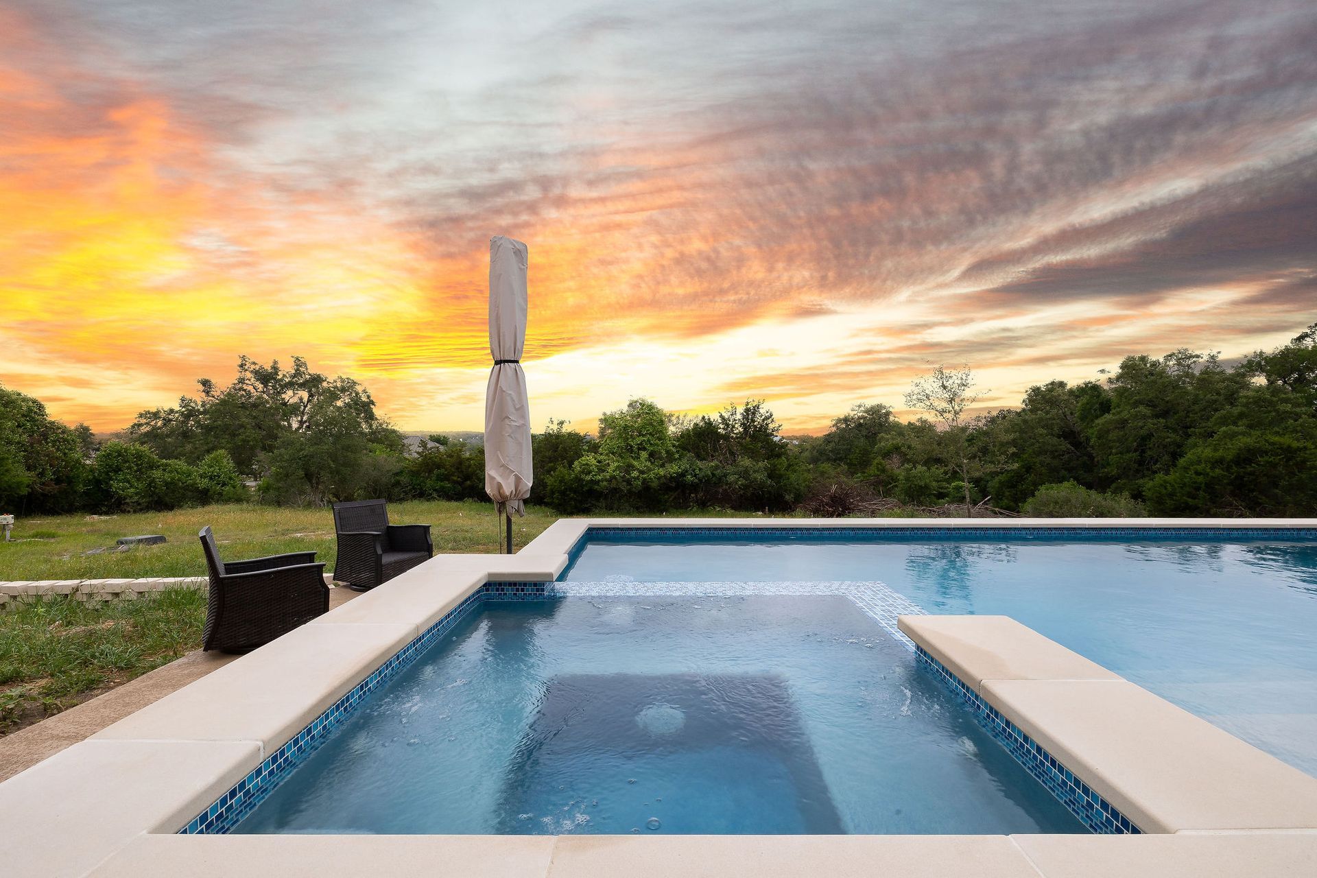 Backyard pool at sunset with a hot tub, lounge chairs, and an umbrella beside a glowing sky