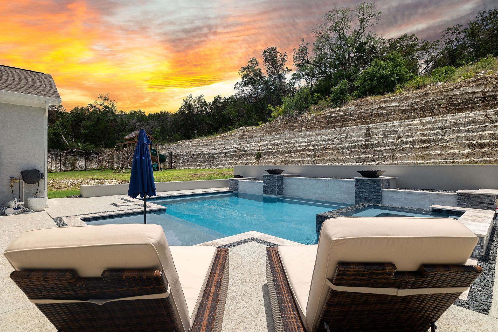 Backyard pool with lounge chairs, umbrella, and sunset sky behind a stone wall