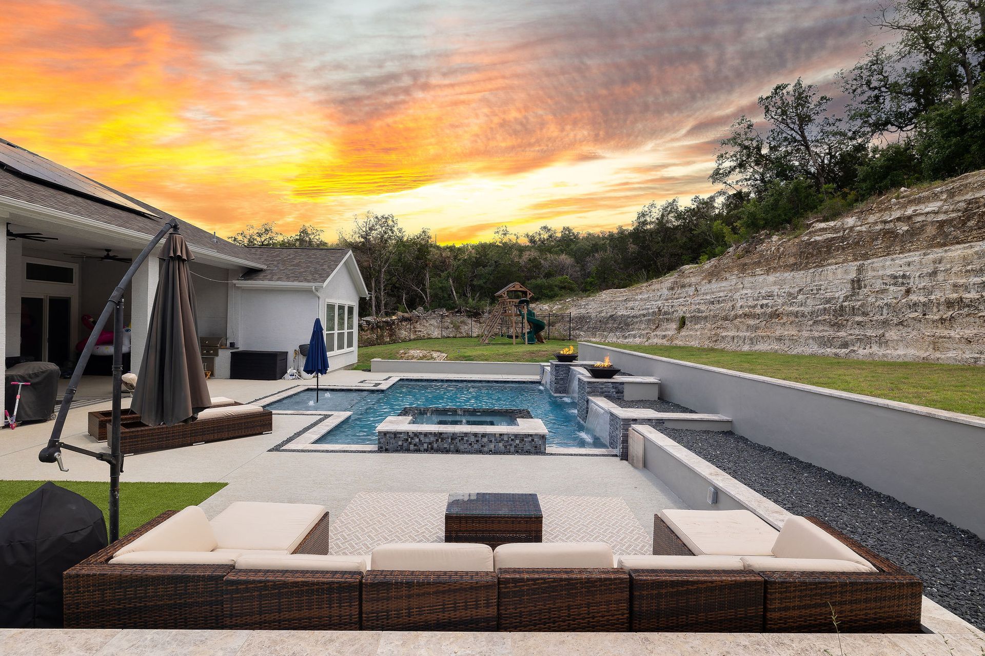 Modern backyard patio with pool, fire pit, lounge seating, and sunset sky behind a house