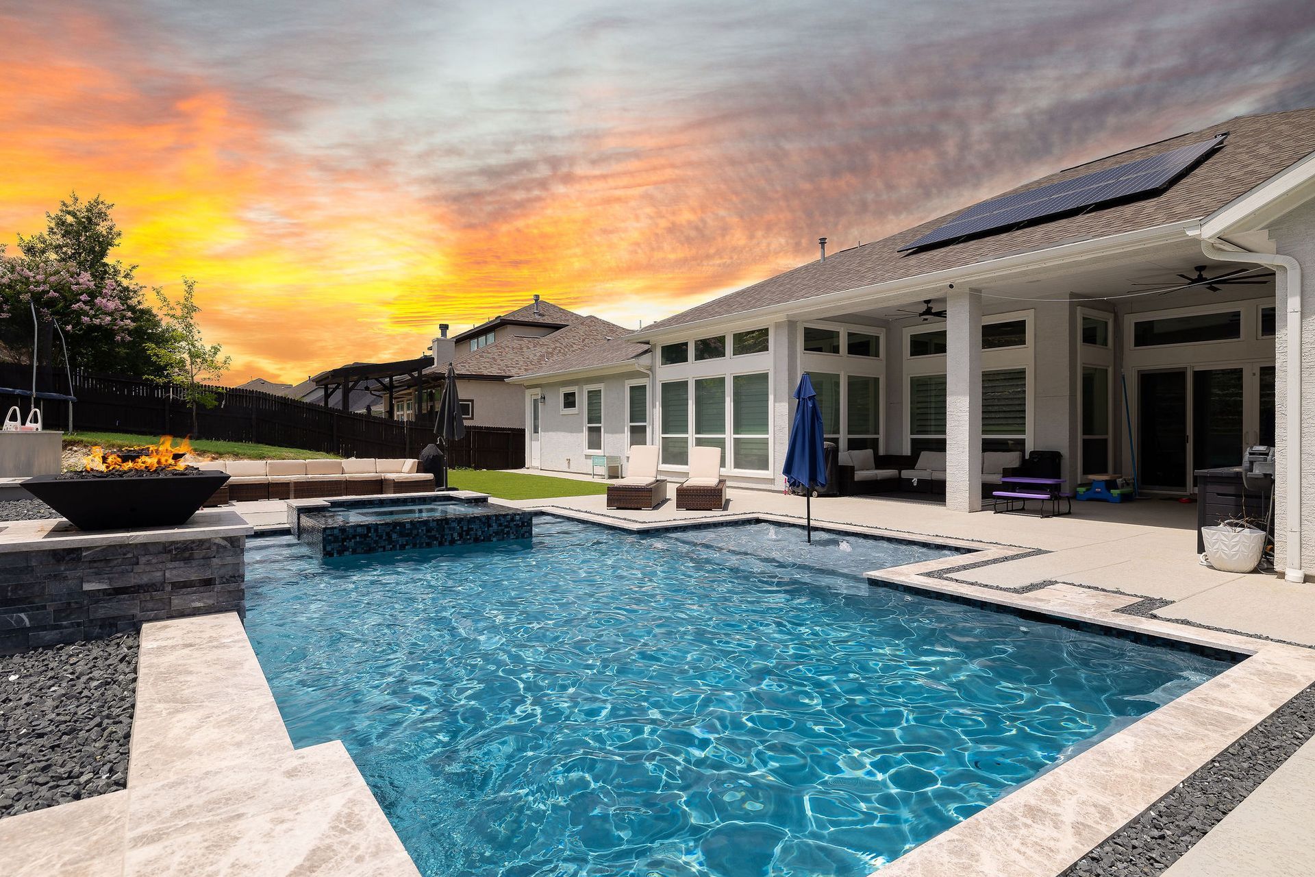 Modern backyard patio with pool, fire pit, lounge seating, and sunset sky behind a house