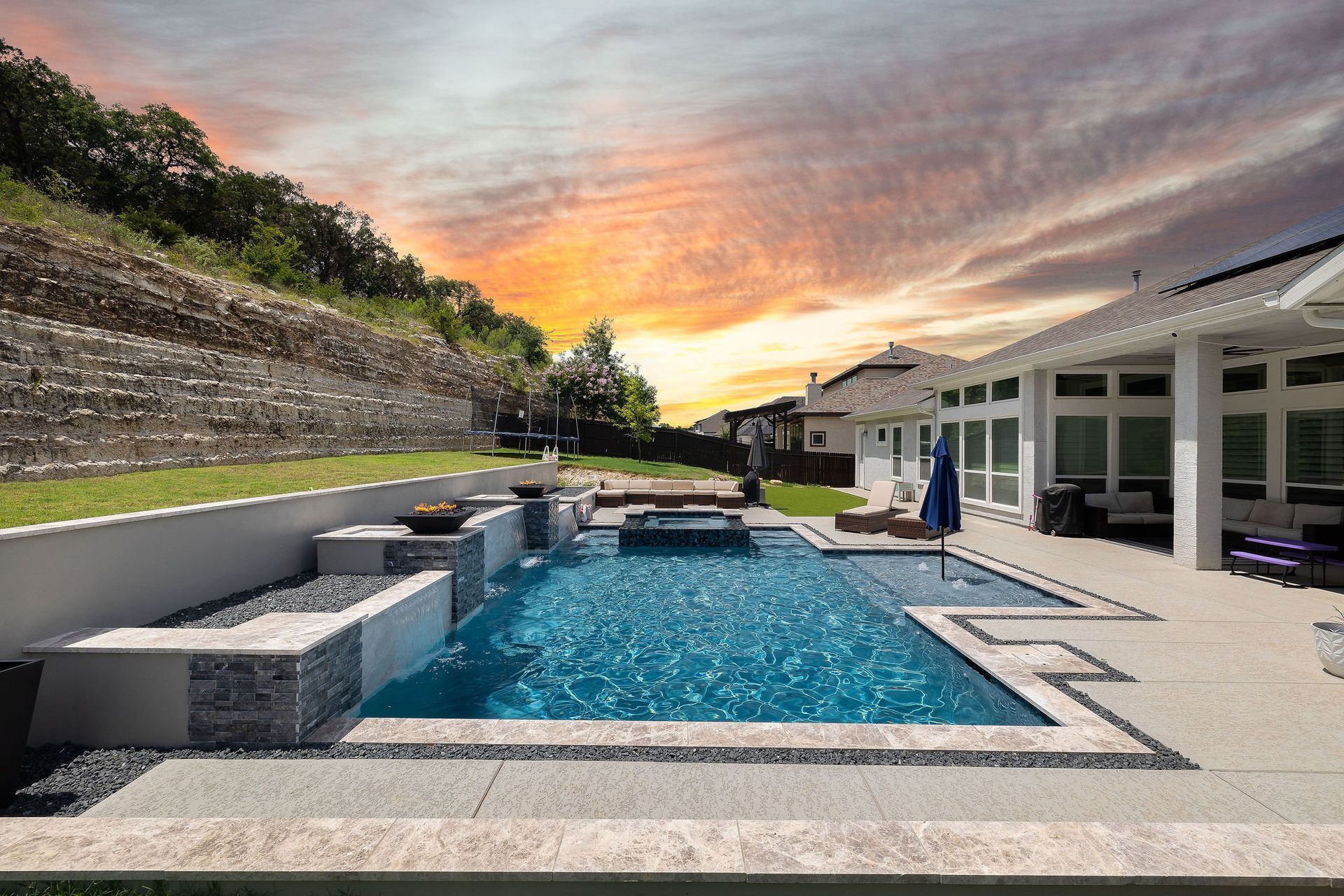 Modern backyard patio with pool, fire pit, lounge seating, and sunset sky behind a house