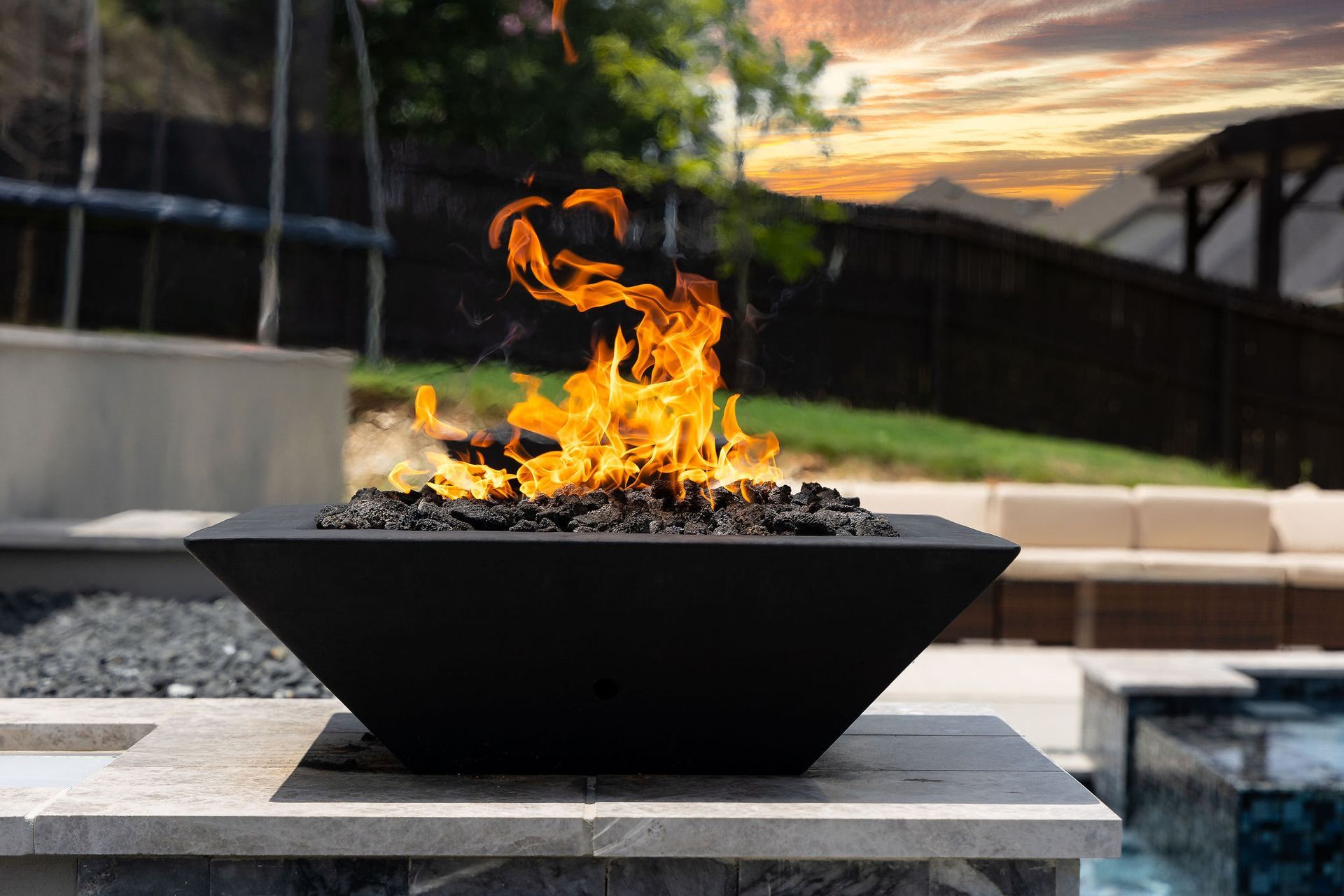 Black fire pit with bright flames on a patio beside a pool at sunset