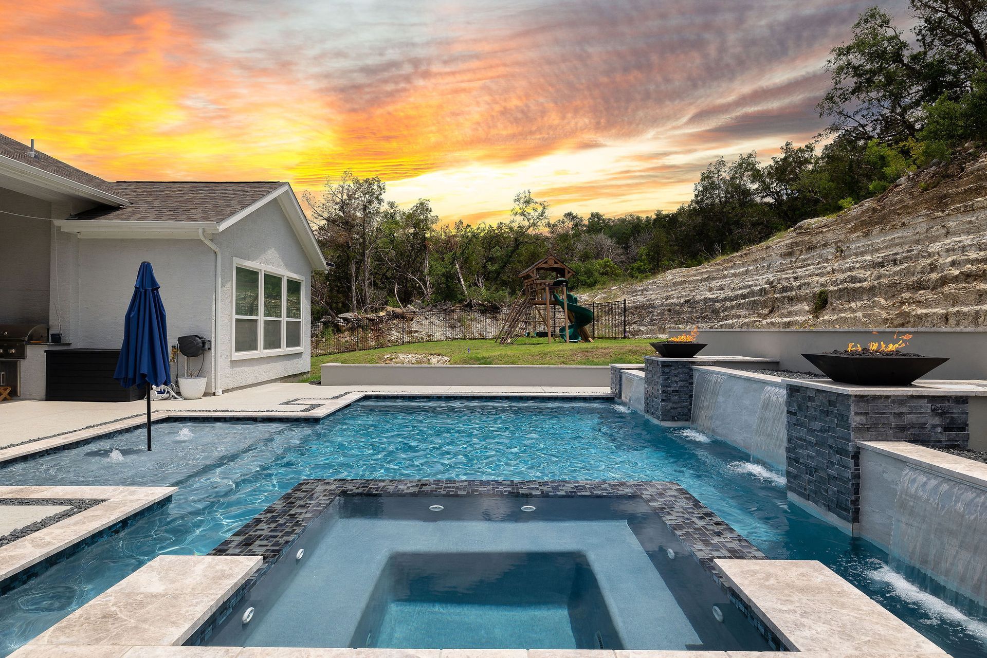 Backyard pool with lounge chairs, umbrella, and sunset sky behind a stone wall