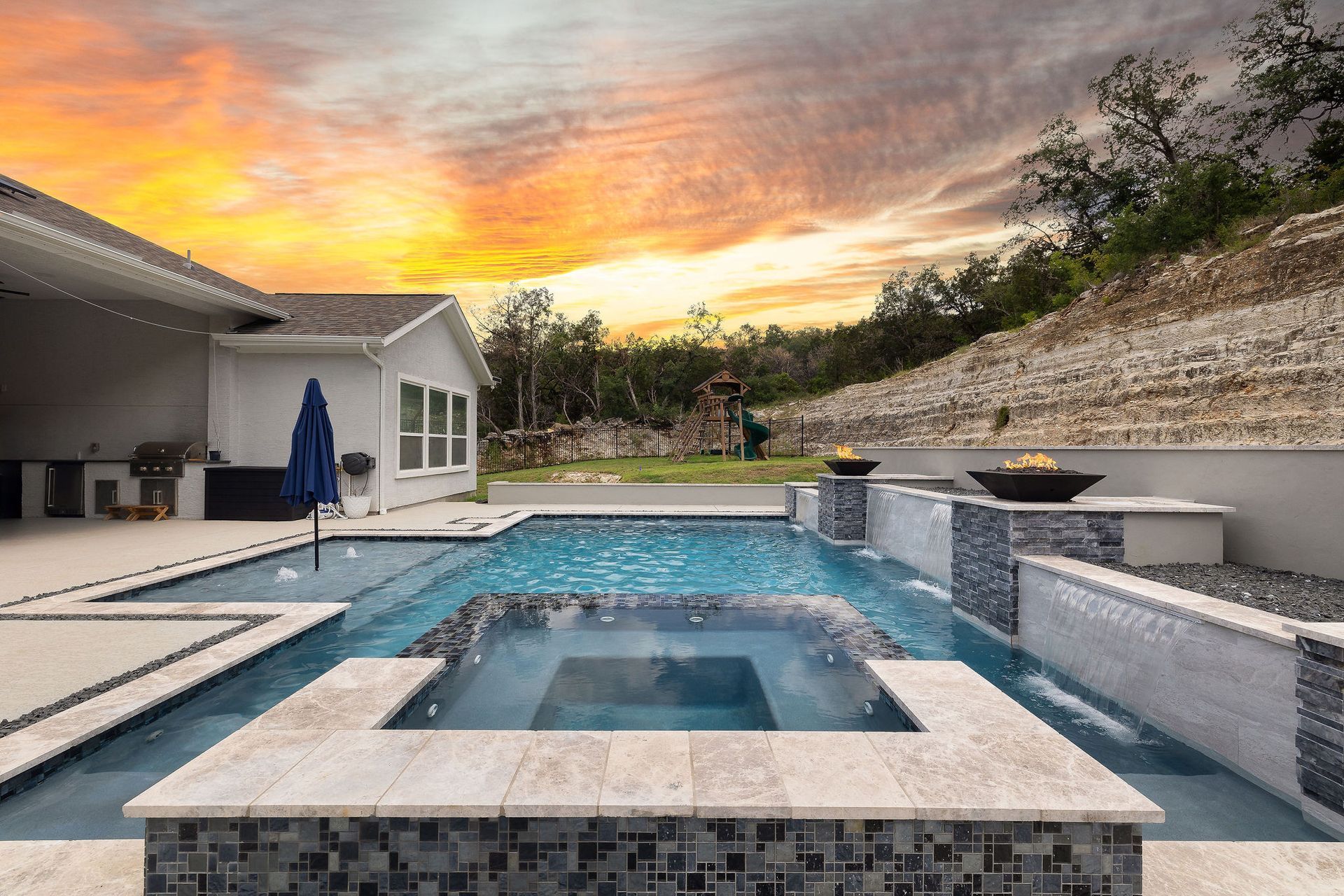 Backyard pool with lounge chairs, umbrella, and sunset sky behind a stone wall