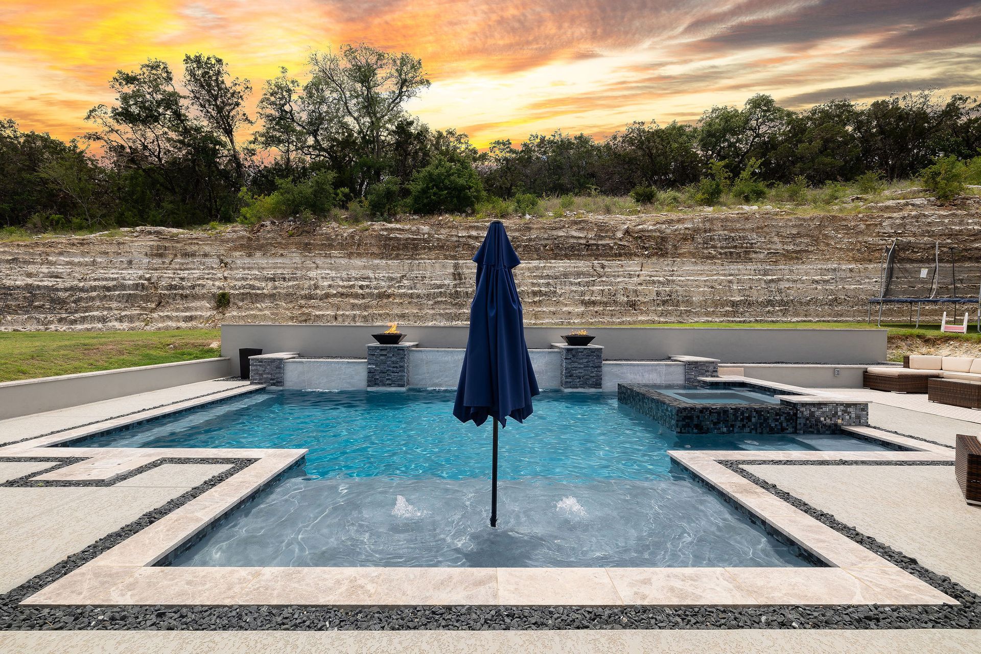 Backyard pool with lounge chairs, umbrella, and sunset sky behind a stone wall
