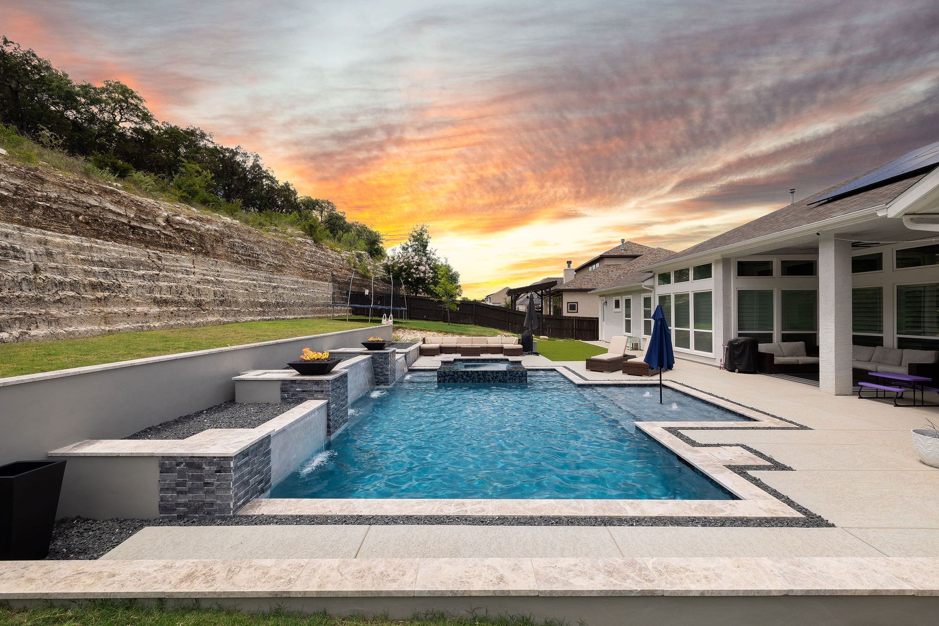 Backyard pool with lounge chairs, umbrella, and sunset sky behind a stone wall