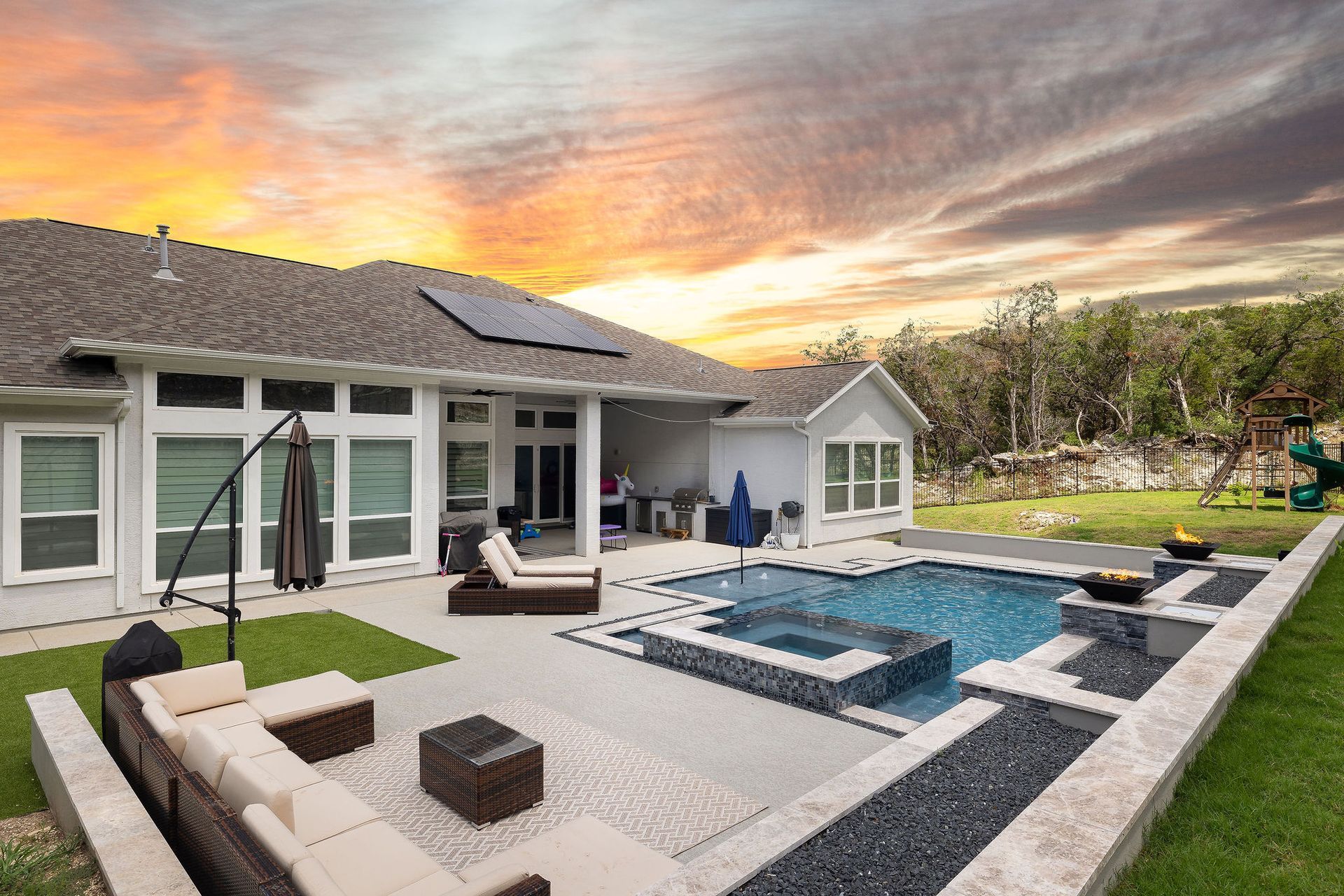 Modern backyard patio with pool, fire pit, lounge seating, and sunset sky behind a house