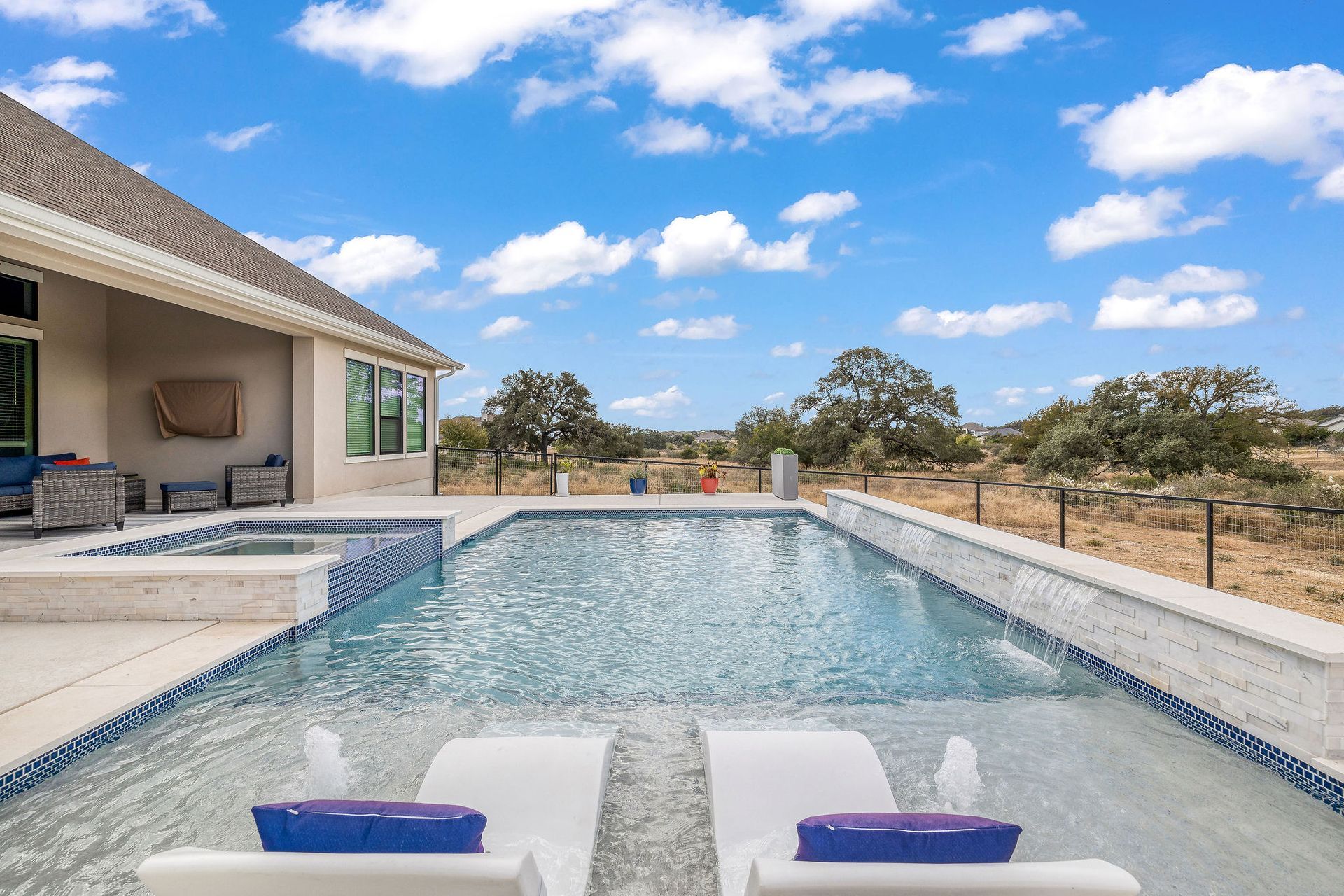 Modern backyard pool beside a house, with lounge chairs, blue sky, and dry open land beyond