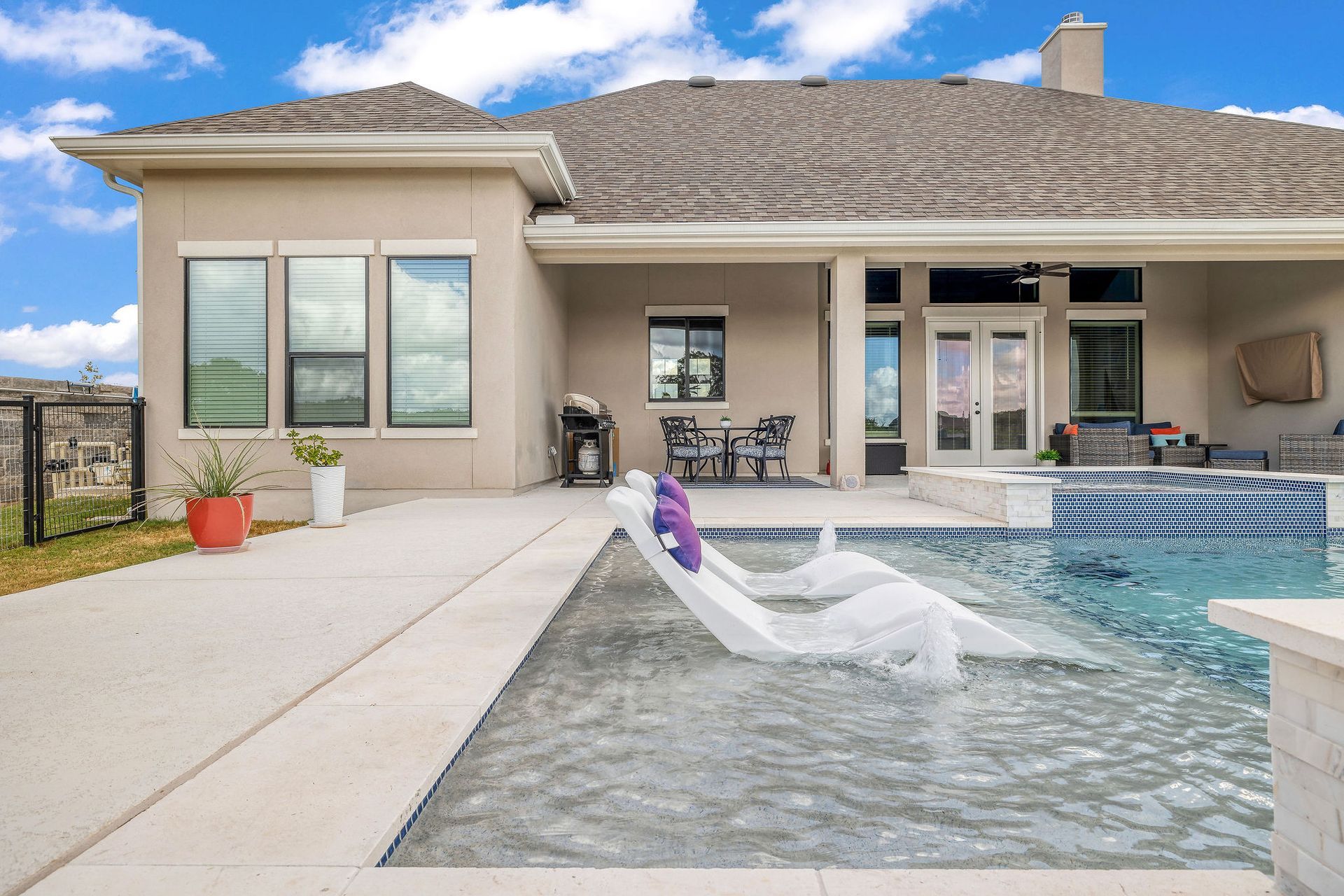 Patio and pool with lounge chair outside a beige house, overlooking a blue swimming pool and covered patio.