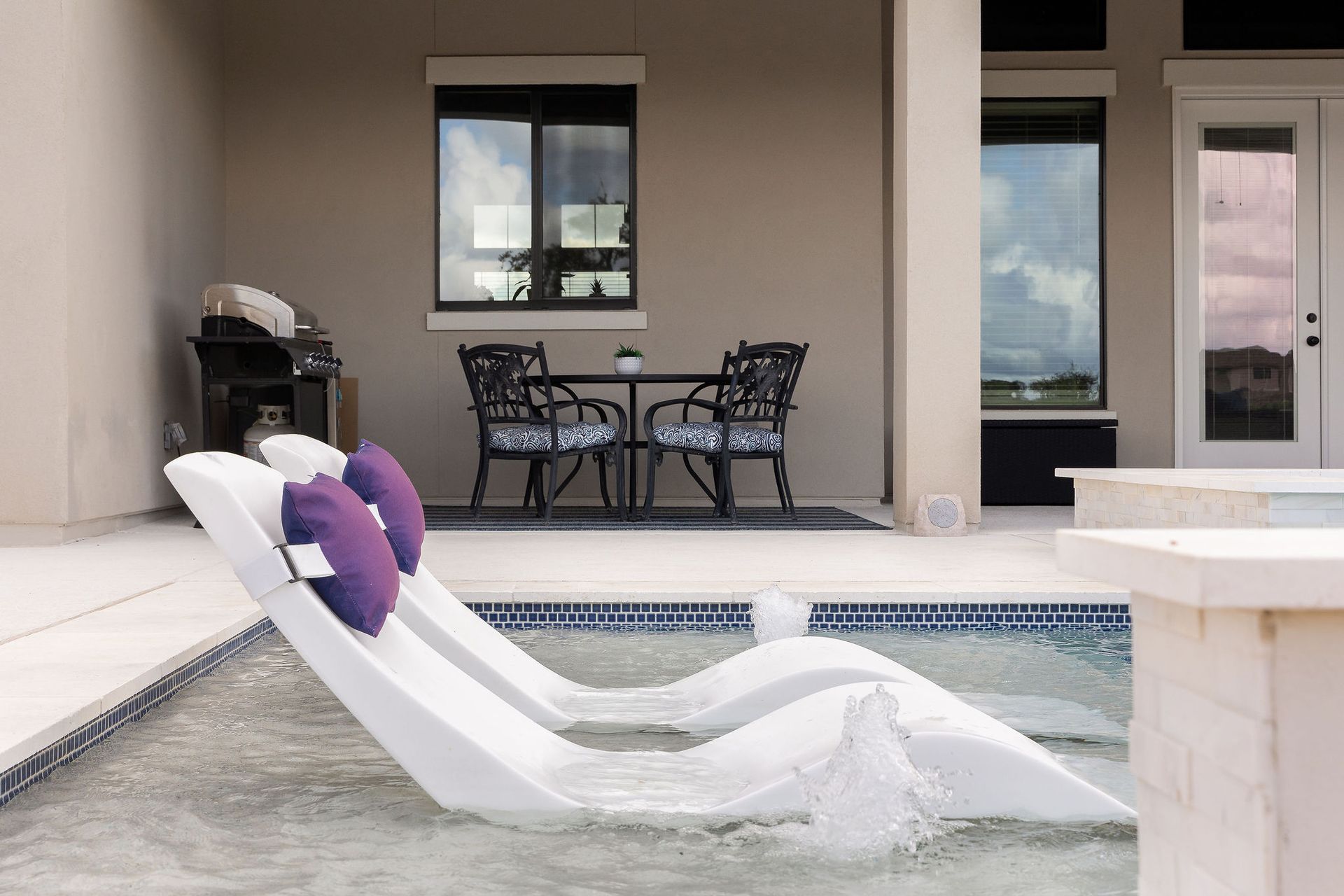 White lounge chair with purple pillows beside a backyard pool and patio seating area