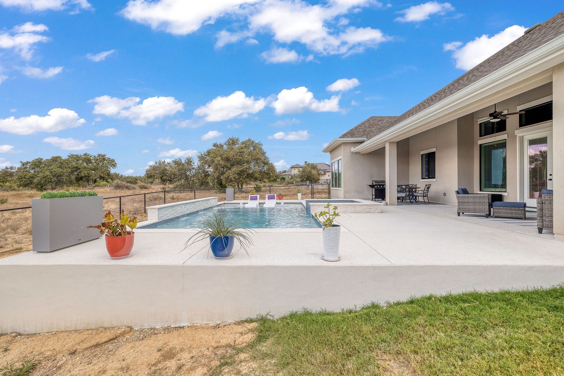 Patio with a pool, potted plants, and lounge chairs beside a modern house under a blue sky