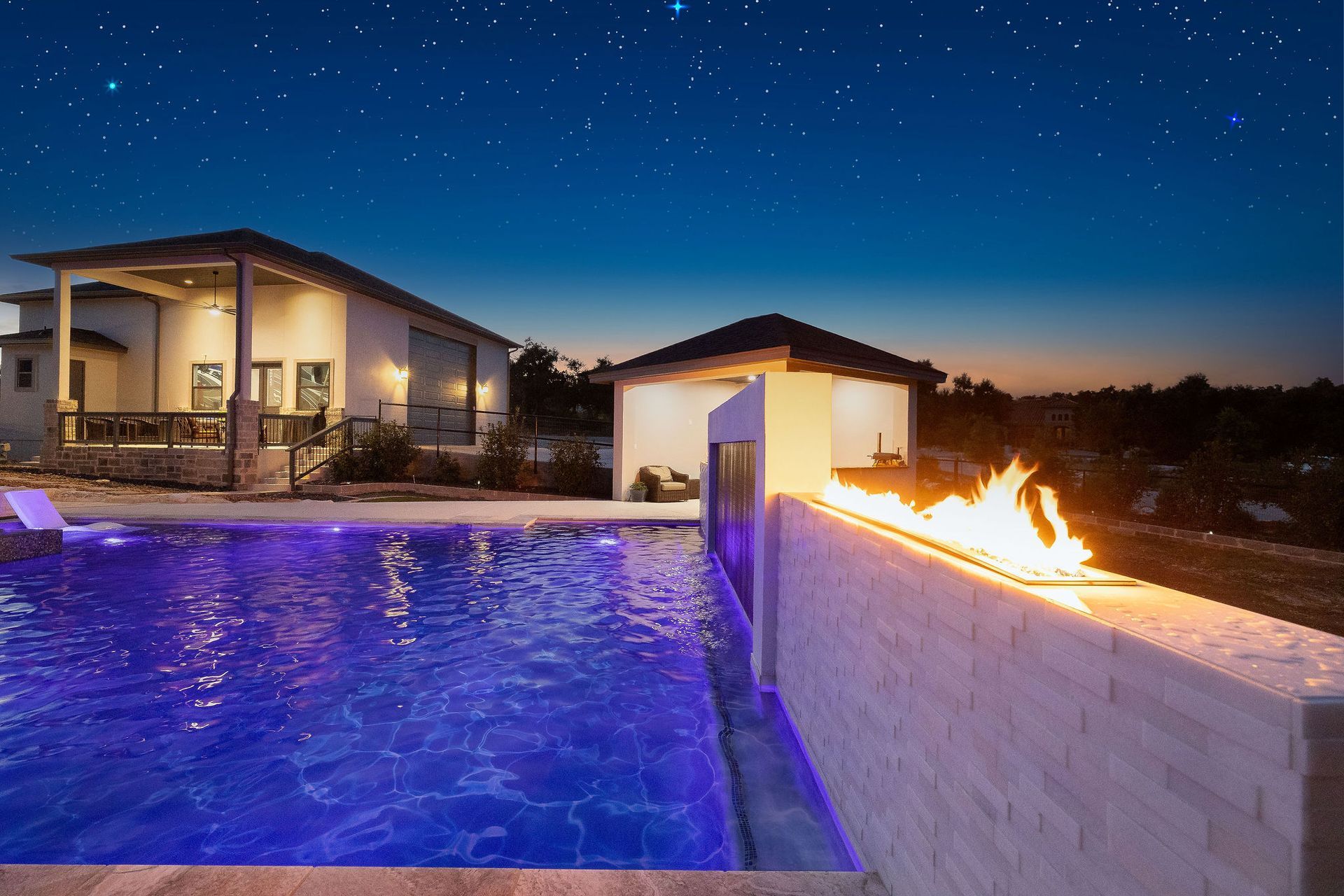 Modern poolside patio at night with a fire wall, lit house, and starry sky