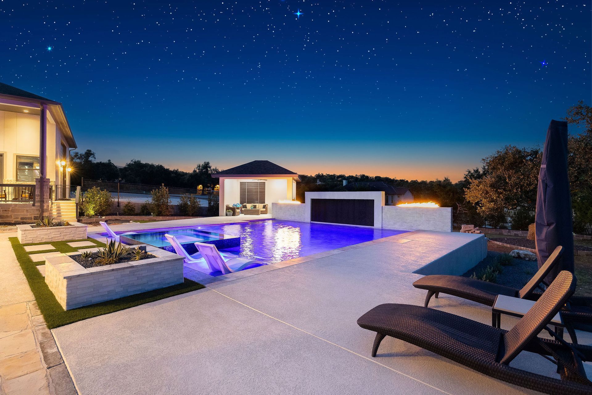 Modern poolside patio at night with a fire wall, lit house, and starry sky
