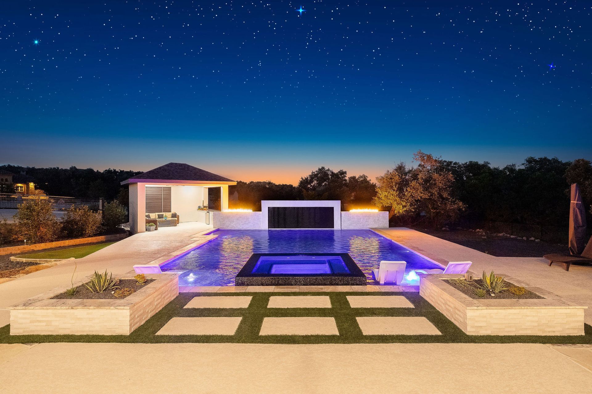 Modern poolside patio at night with a fire wall, lit house, and starry sky