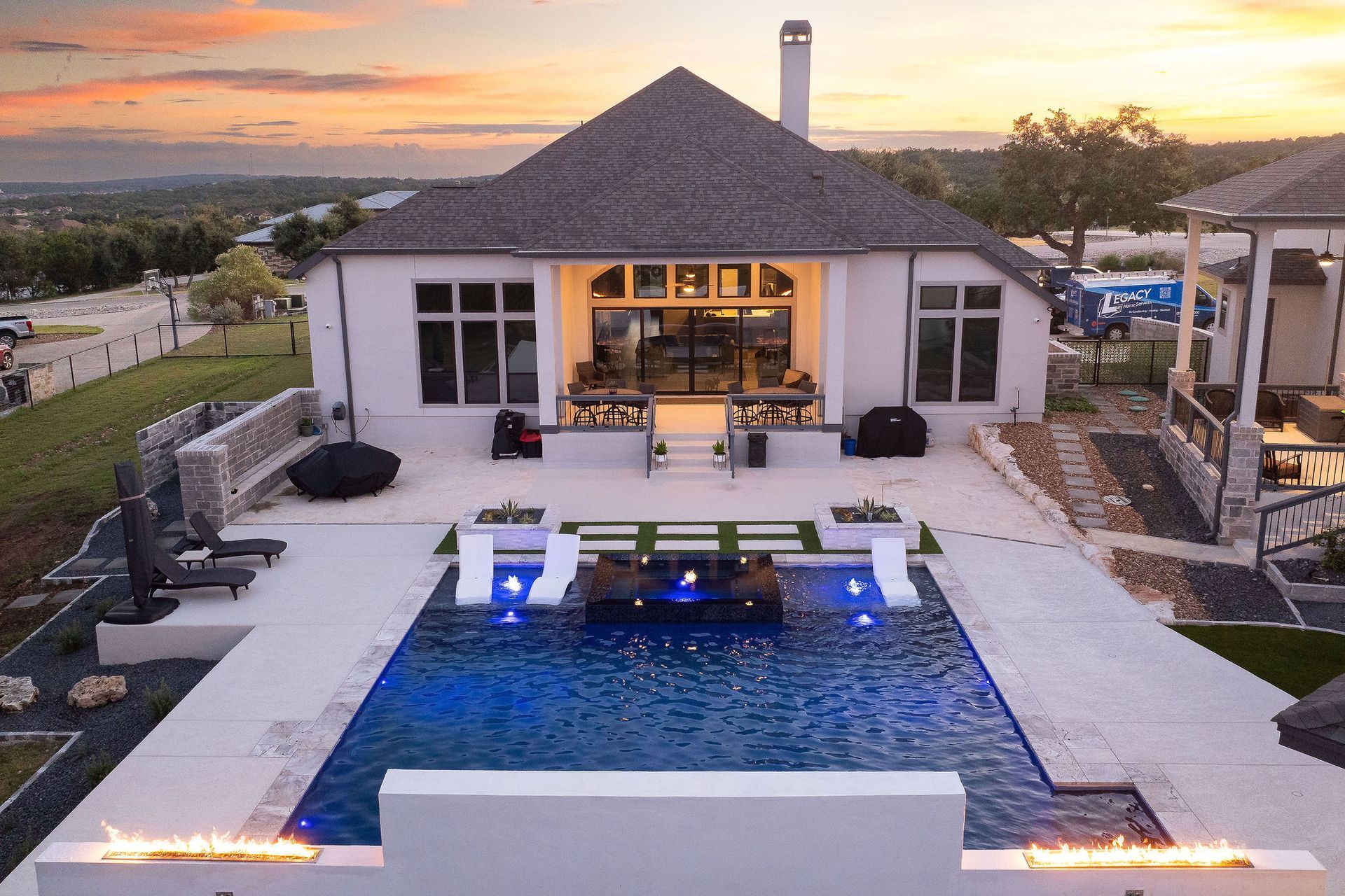 Modern poolside patio at night with a fire wall, lit house, and starry sky