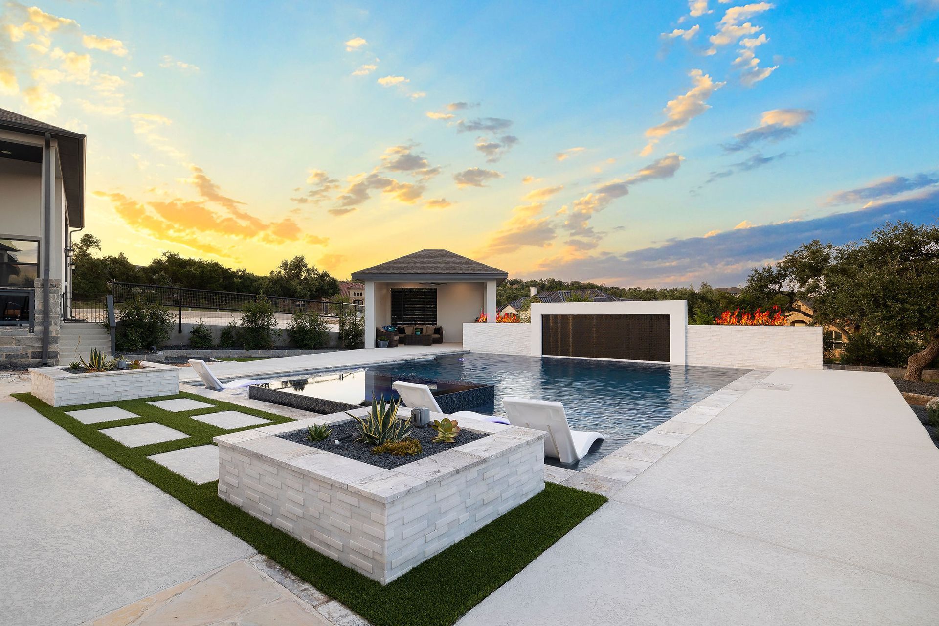 Modern backyard with a pool, white patio, fire pit, and a small pavilion at sunset