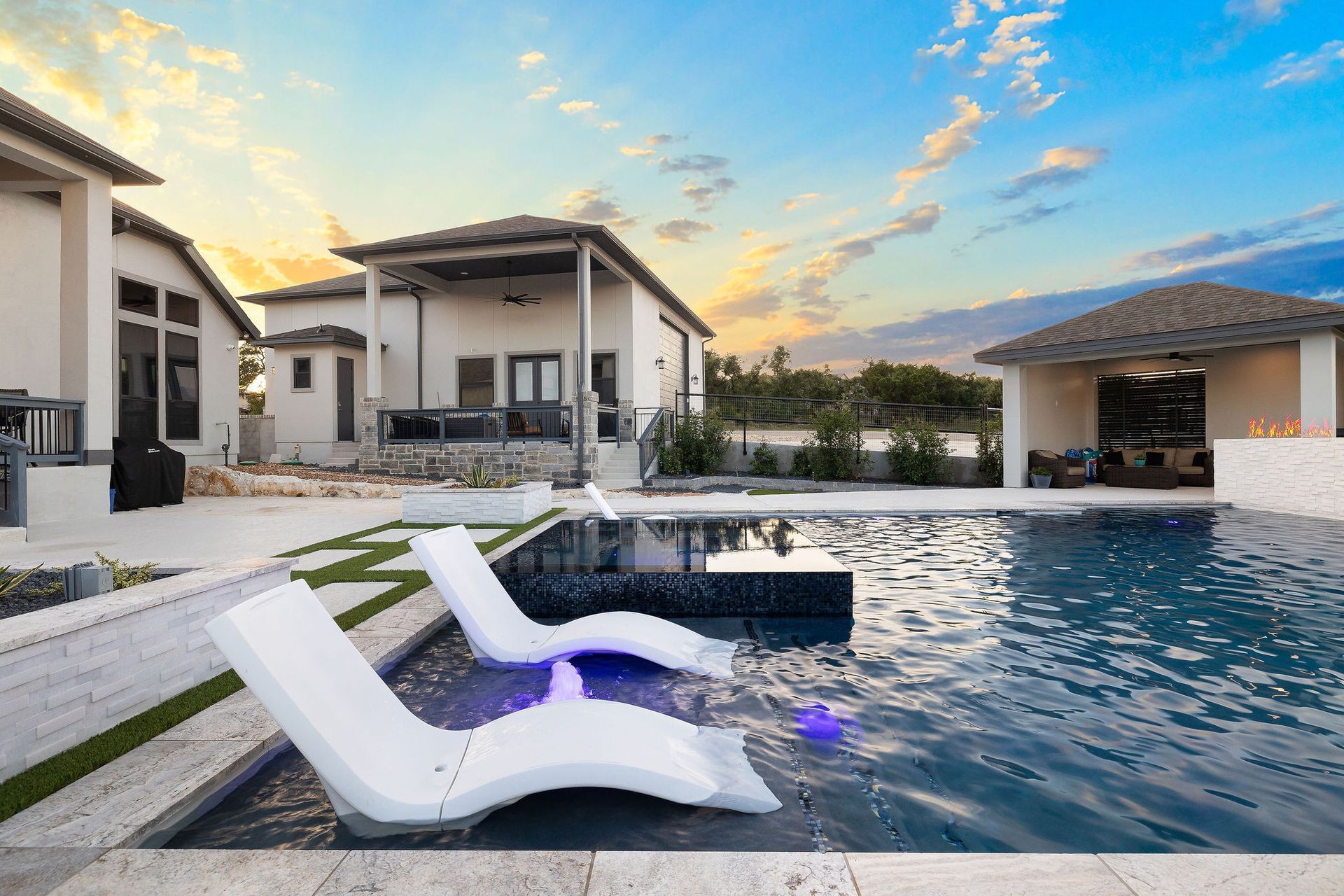 Modern villa pool at sunset with white loungers, calm water, and a glowing sky