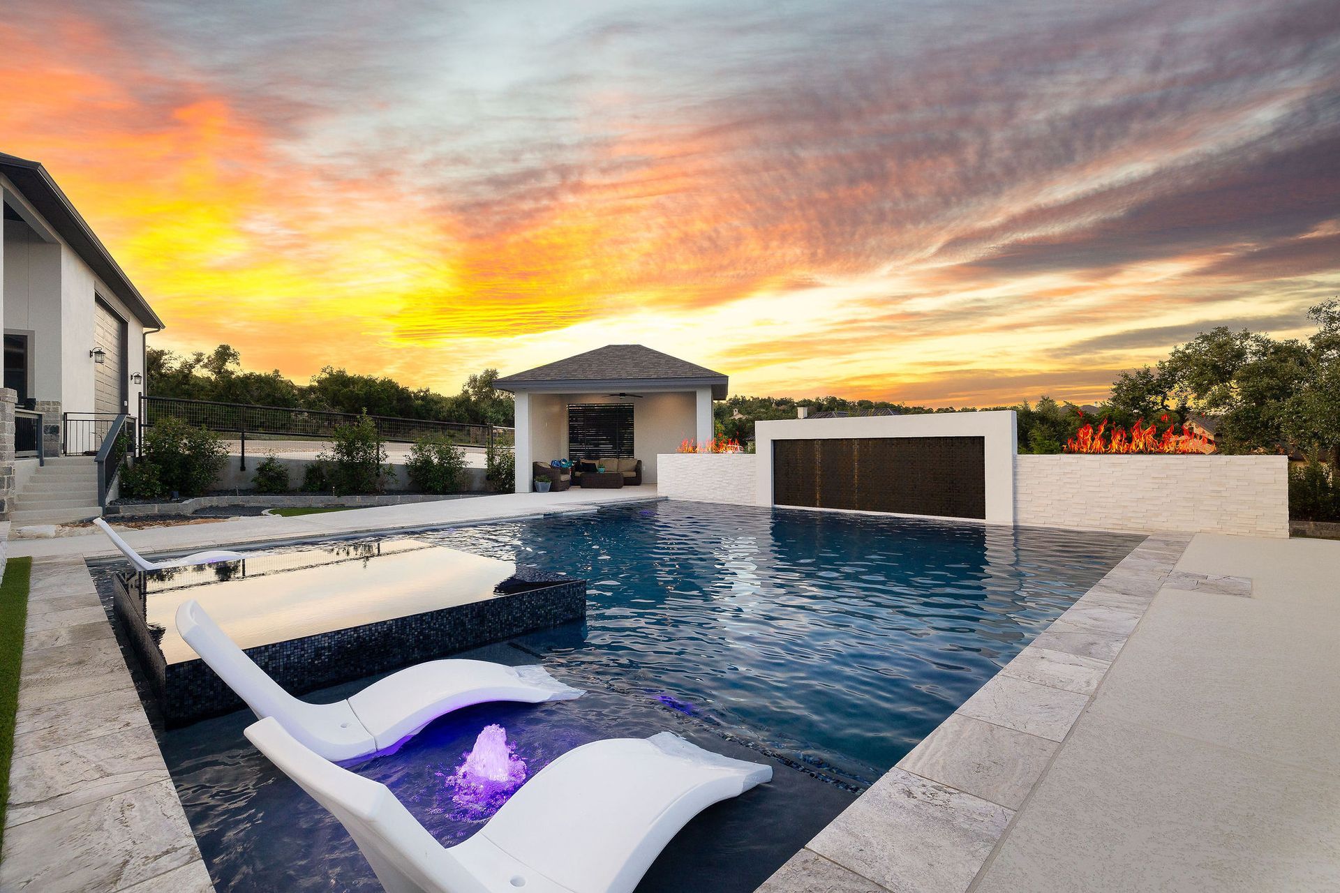 Modern backyard pool at sunset with lounge chairs and a lit spa, framed by a sleek patio and dramatic sky