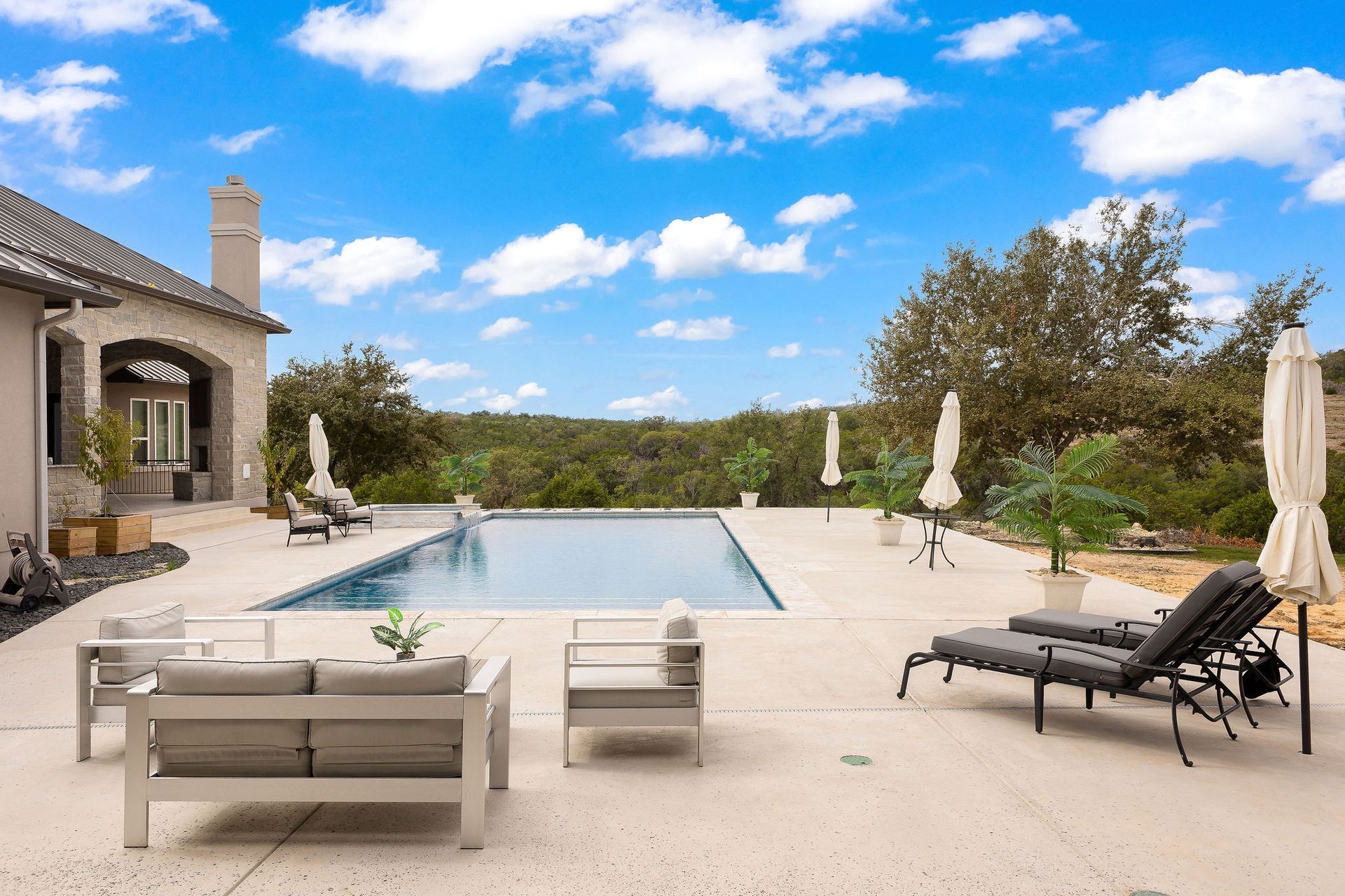 Modern backyard patio with a pool, lounge chairs, umbrellas, and stone house under a blue sky