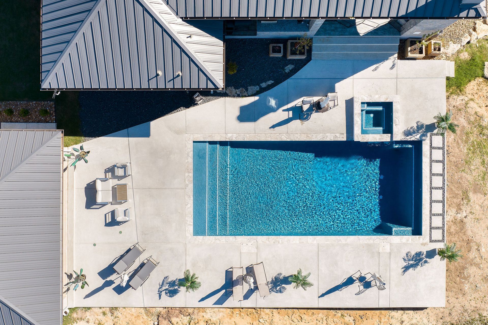 Aerial view of a backyard with a rectangular blue swimming pool and lounge chairs on a stone patio.