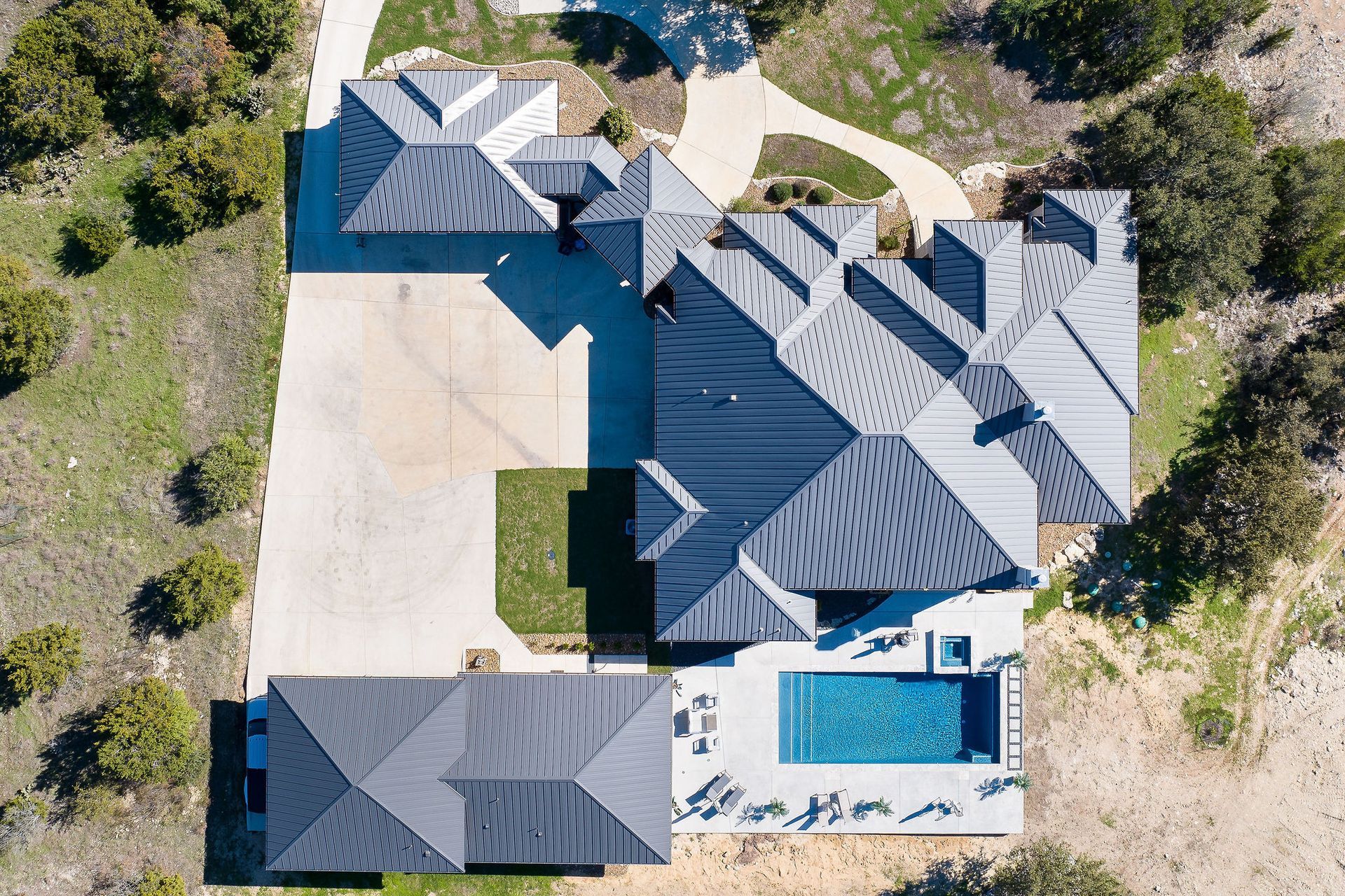 Aerial view of a large house with a blue roof, driveway, patio, and swimming pool surrounded by trees.