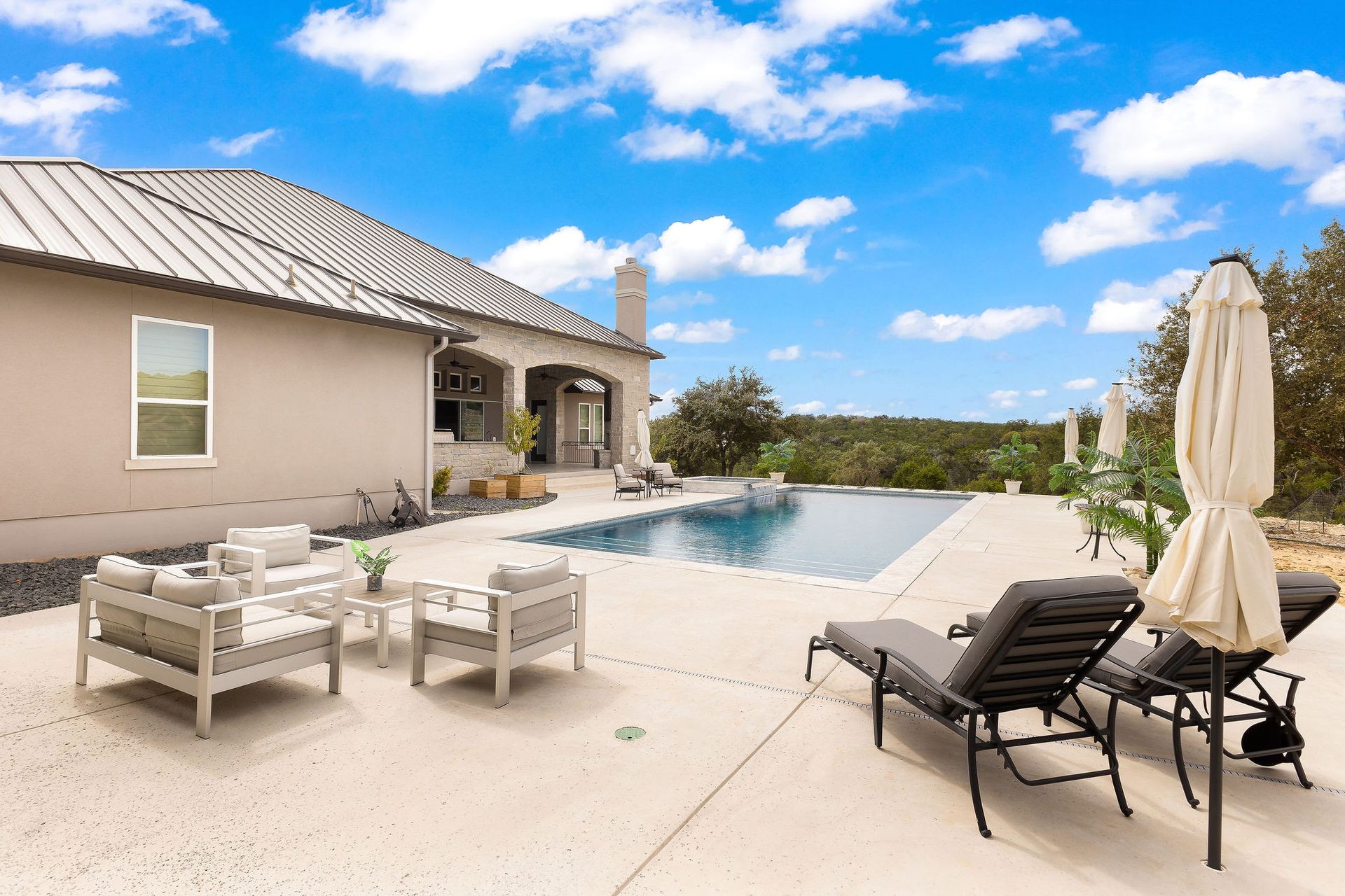 Poolside patio with outdoor seating beside a beige house under a bright blue sky