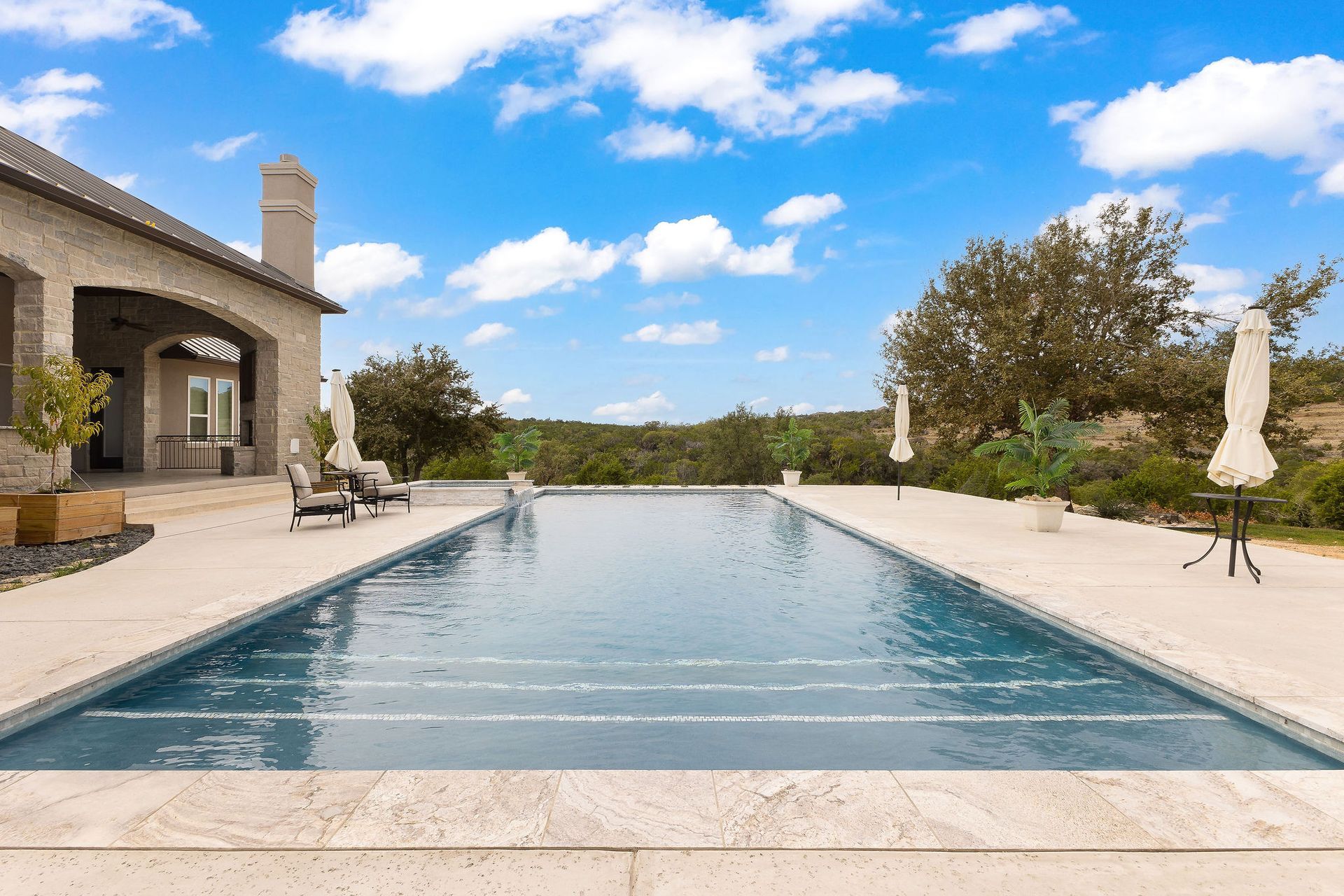 Infinity pool beside a stone villa under a blue sky with lounge chairs and trees