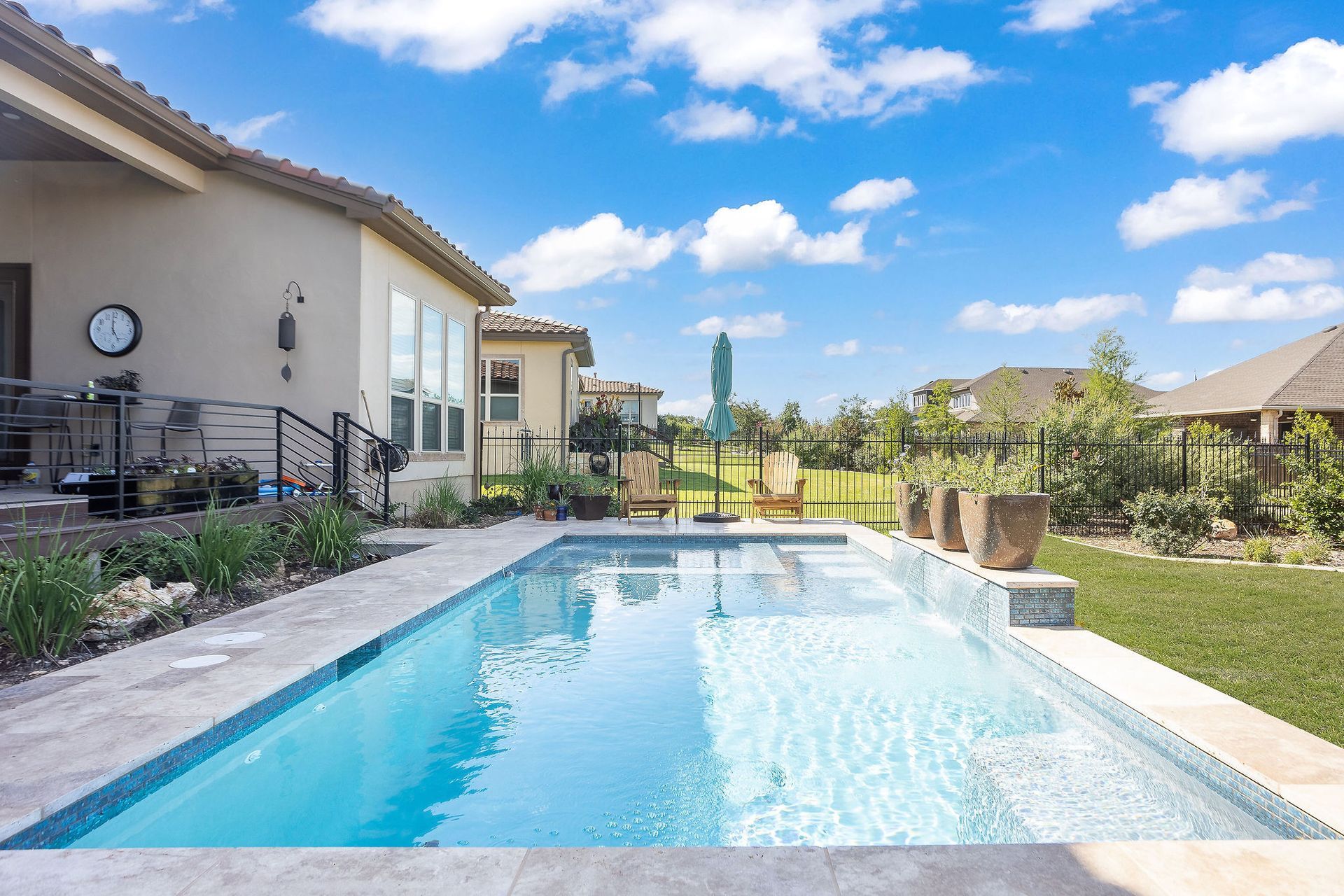 Backyard swimming pool beside a house, with patio, fence, and blue sky with clouds