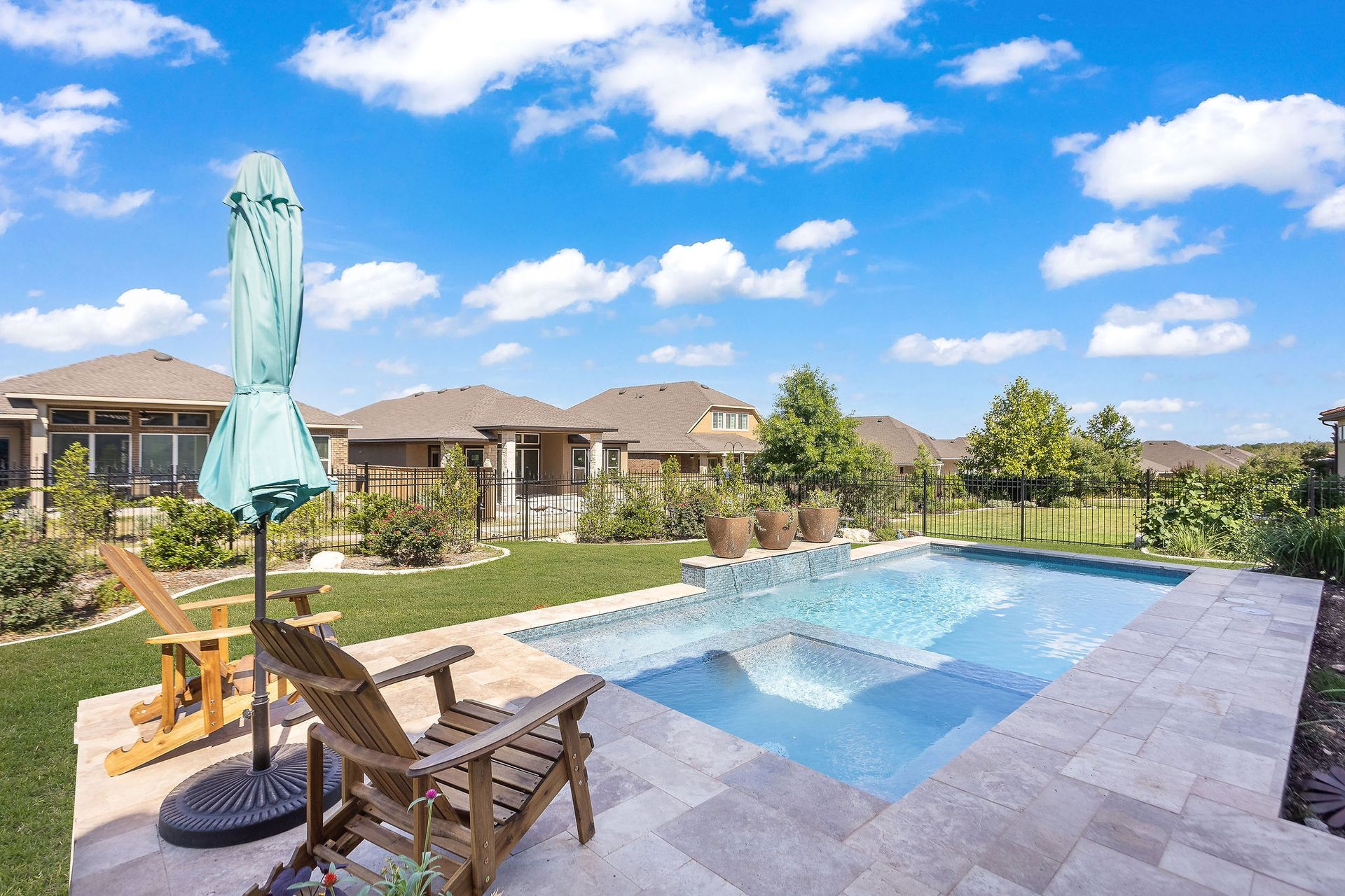 Patio view of a backyard pool with lounge chairs, umbrella, and modern homes under a blue sky