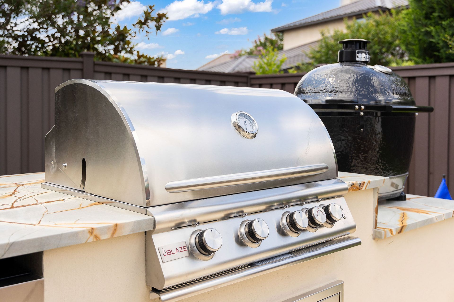 Stainless steel gas grill beside a black ceramic smoker on a sunlit patio with a wooden fence.