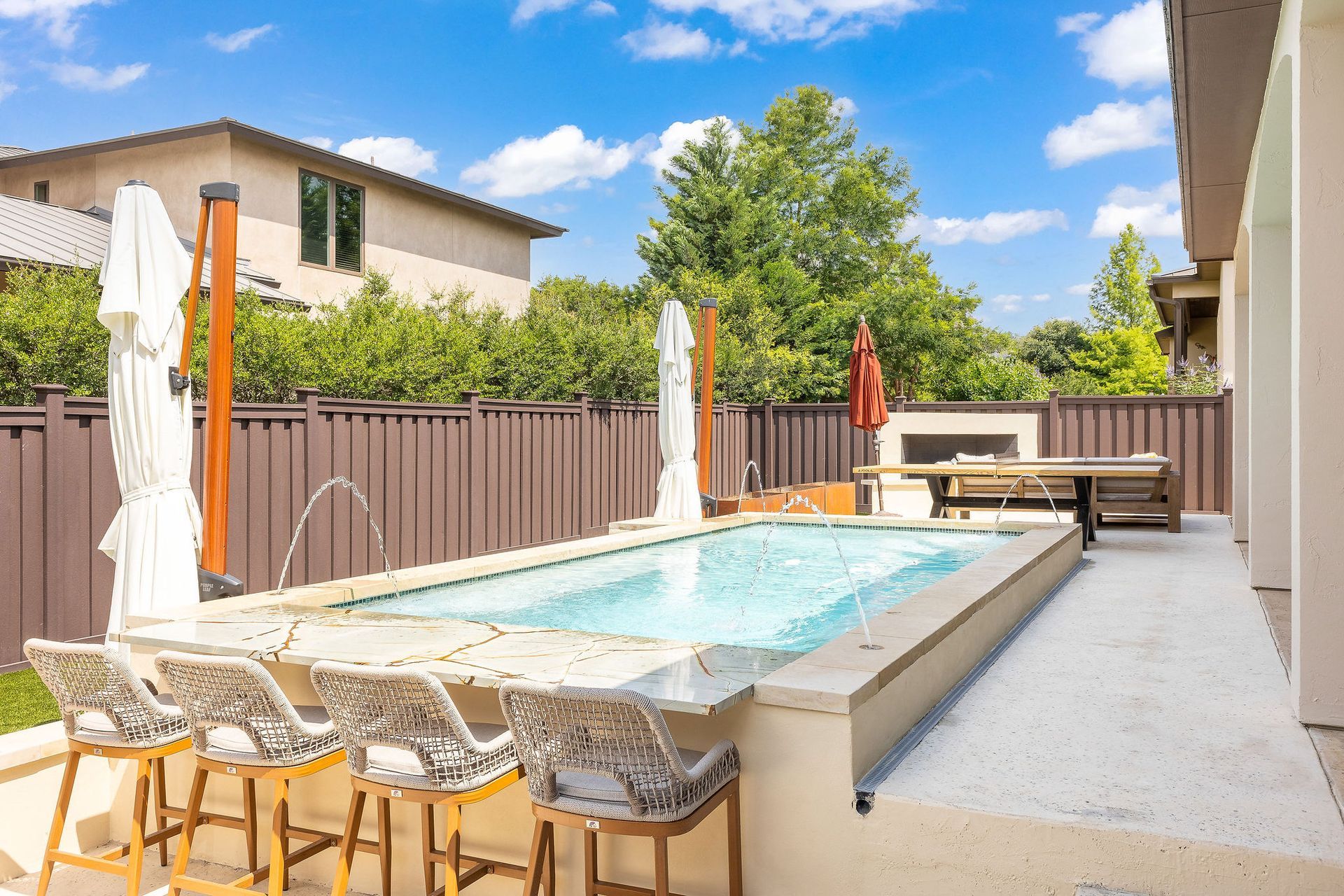 Backyard pool with lounge chairs, umbrellas, and a wooden fence under a bright blue sky