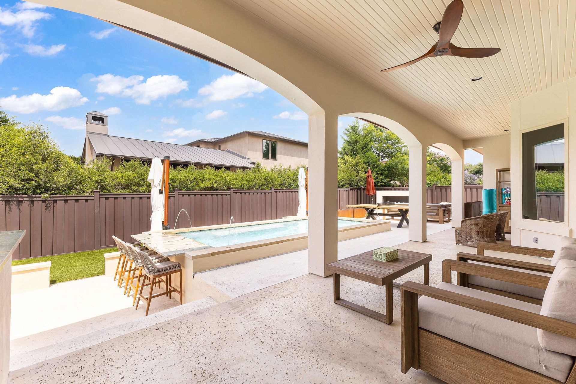 Covered patio with seating overlooking a backyard pool and fence, bright sunny day