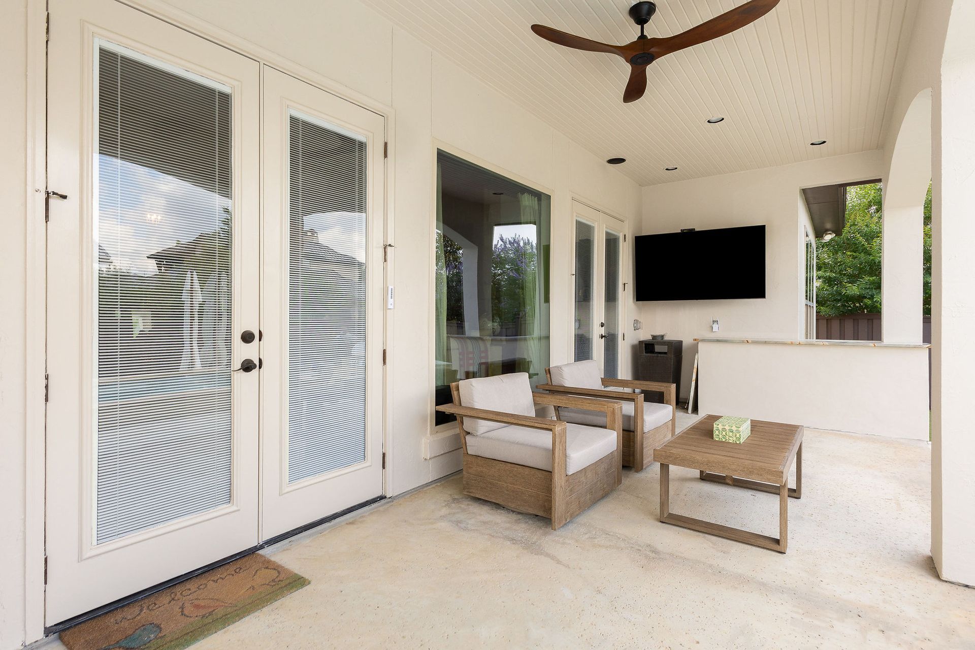 Covered patio with wicker seating, ceiling fan, wall-mounted TV, and white French doors with blinds