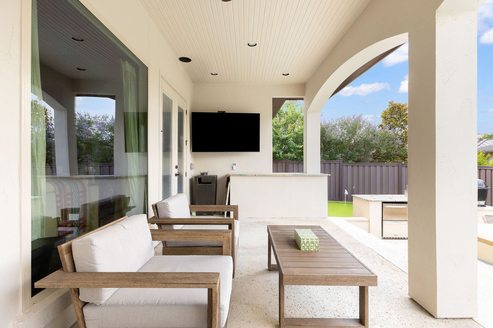 Covered patio with white sofas, wooden coffee table, and wall-mounted TV overlooking a backyard