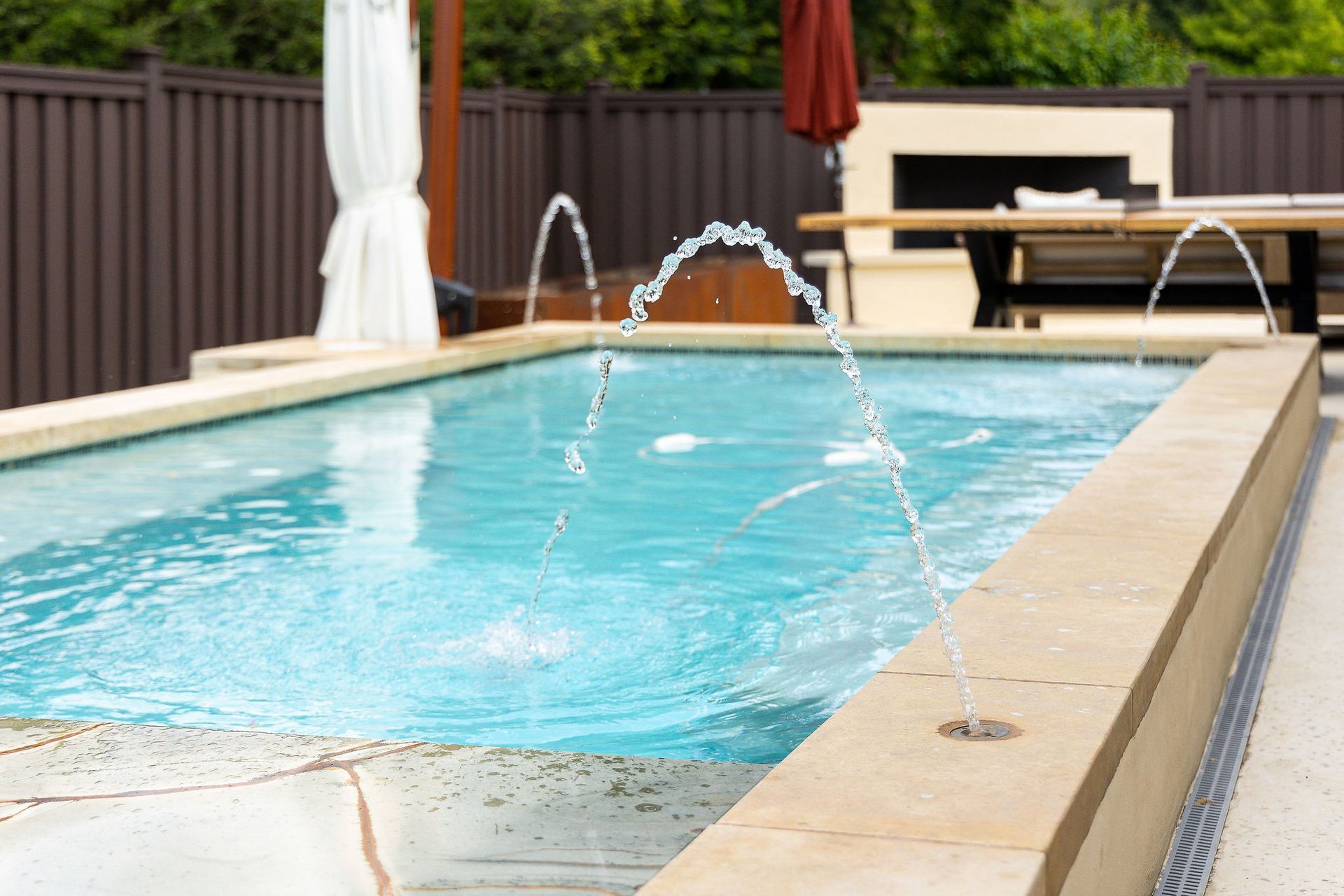 Backyard swimming pool with water jets arcing into clear blue water, bordered by tan coping and a wooden fence