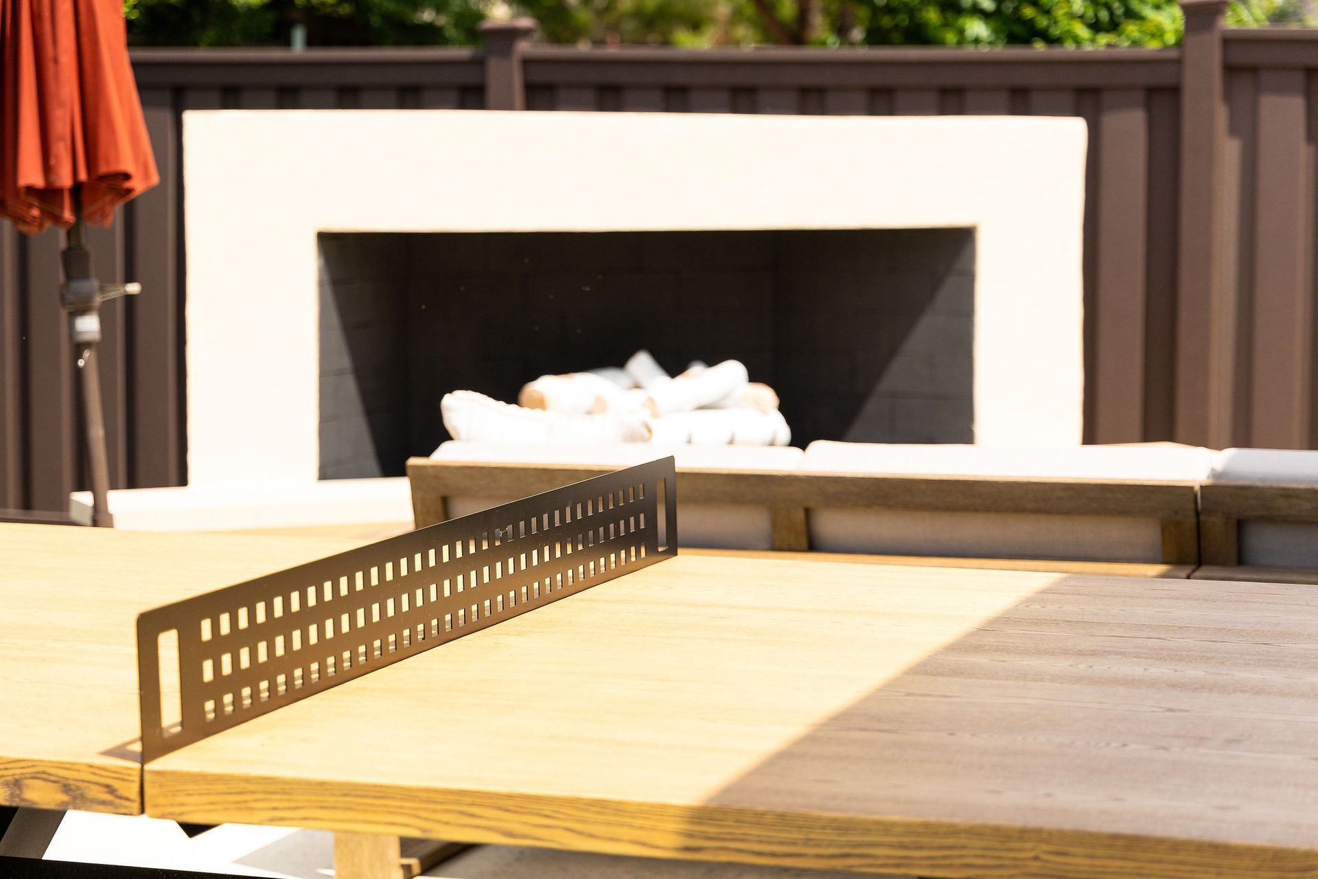 Outdoor wooden table with a built-in ping-pong net on a patio, with a white fire pit in the background
